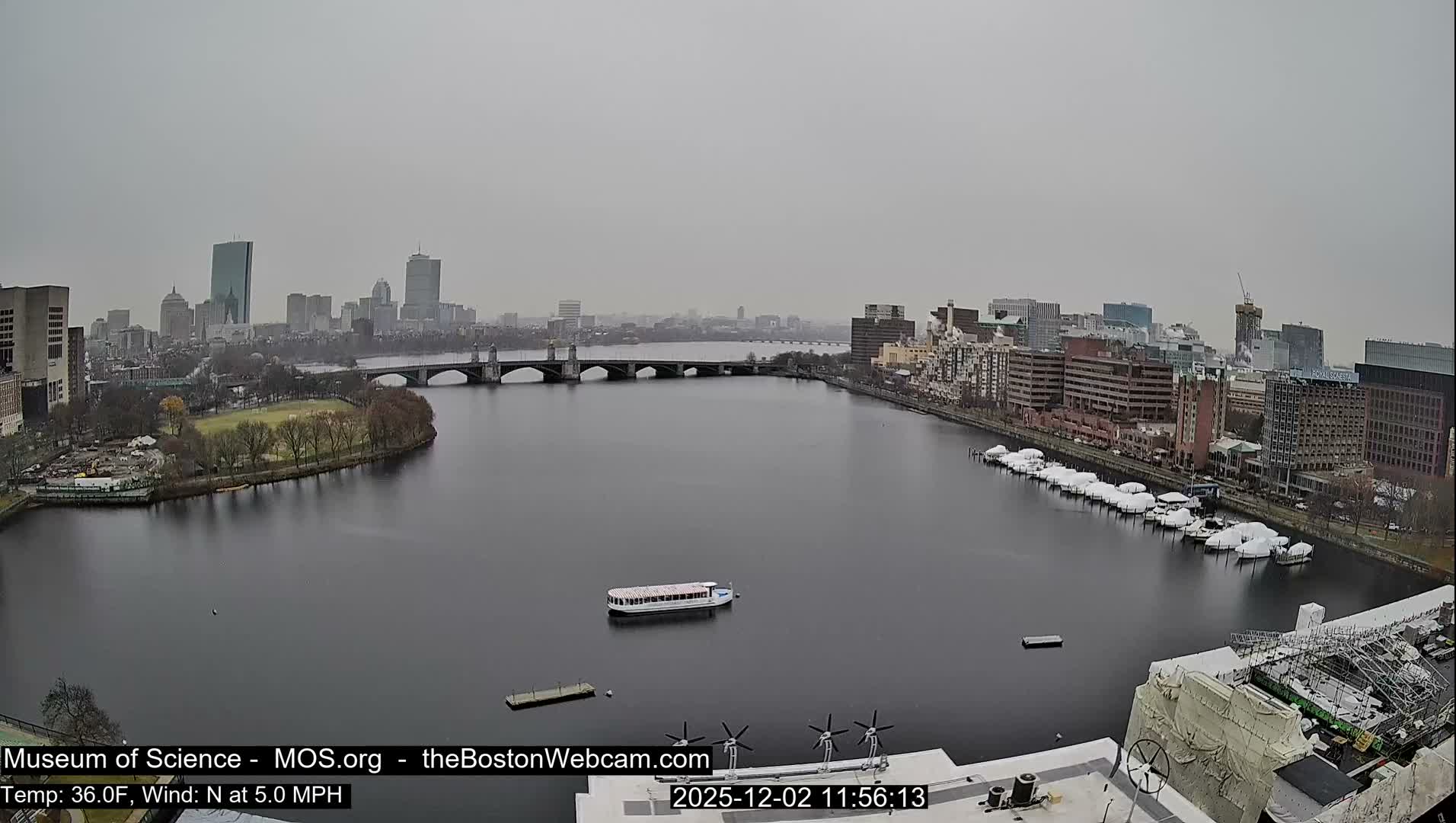 An aerial view reveals a city skyline across a wide river, featuring multiple bridges, a tree-lined island, and numerous winterized boats docked along the bank, all under a uniformly gray and overcast sky.