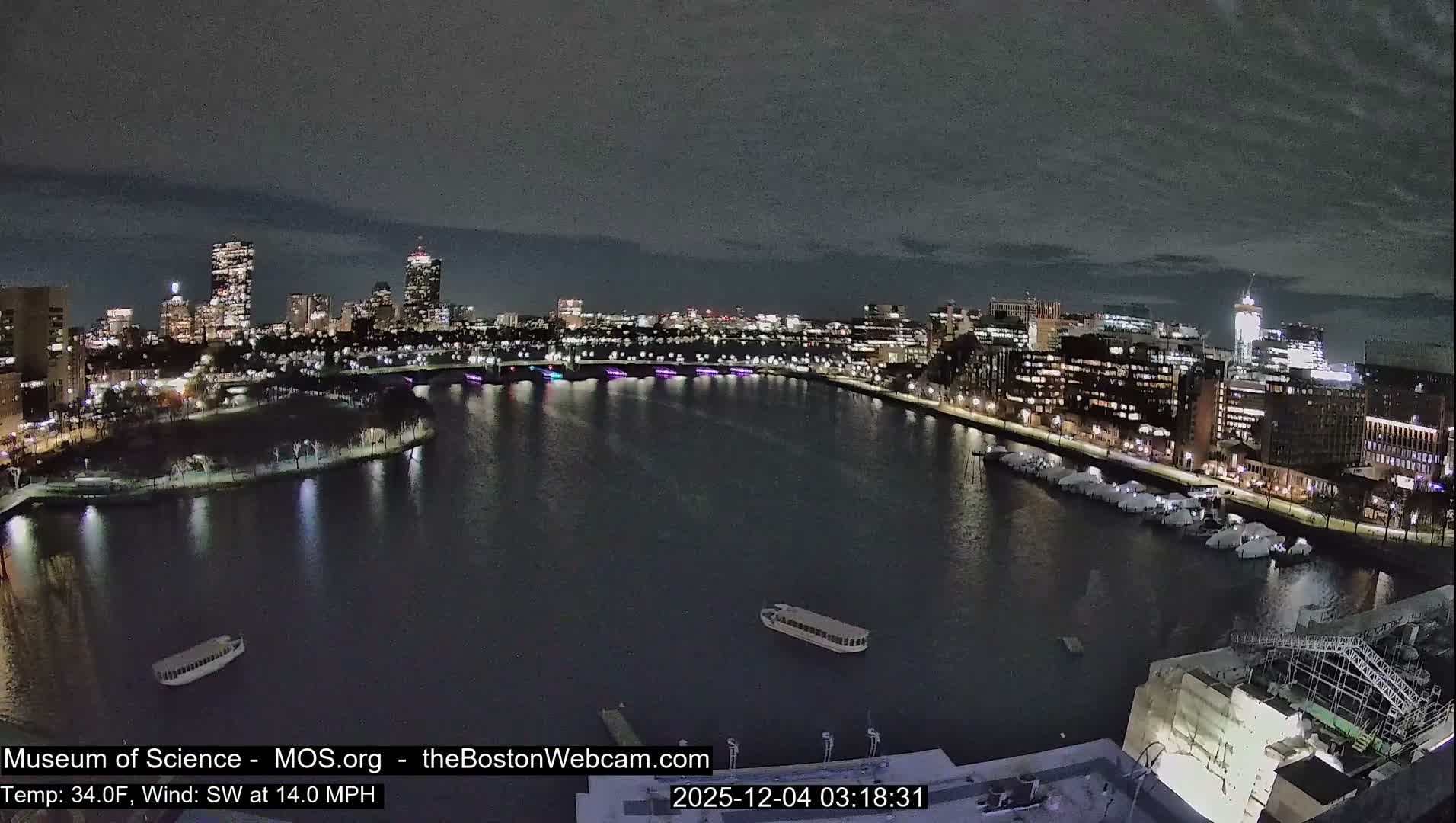 An illuminated cityscape stretches along a wide river or harbor at night, featuring a colorful bridge, numerous buildings with glowing windows, and several boats on the water and docked along the banks, all under a cloudy sky.