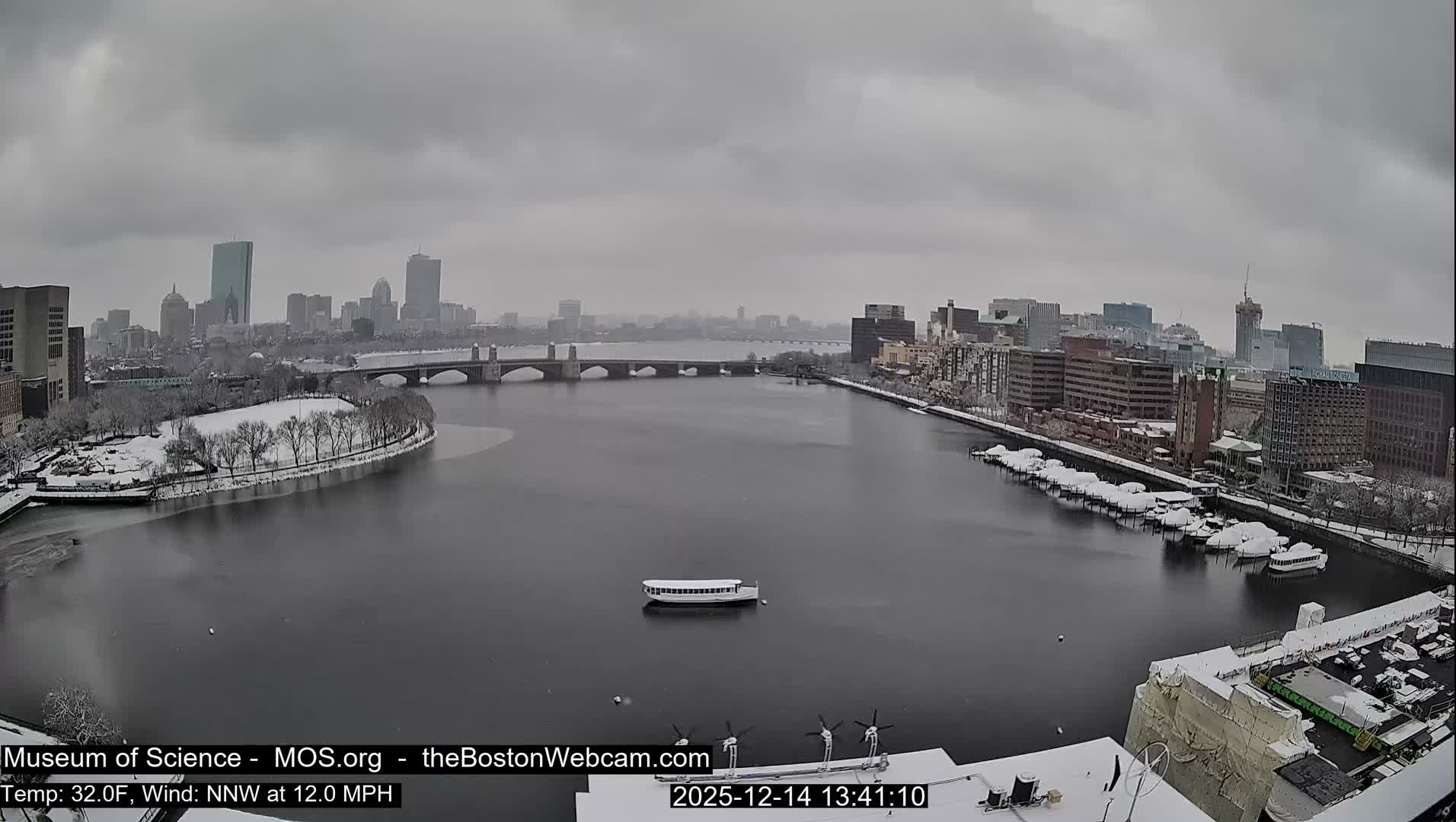 An illuminated cityscape stretches along a wide river or harbor at night, featuring a colorful bridge, numerous buildings with glowing windows, and several boats on the water and docked along the banks, all under a cloudy sky.