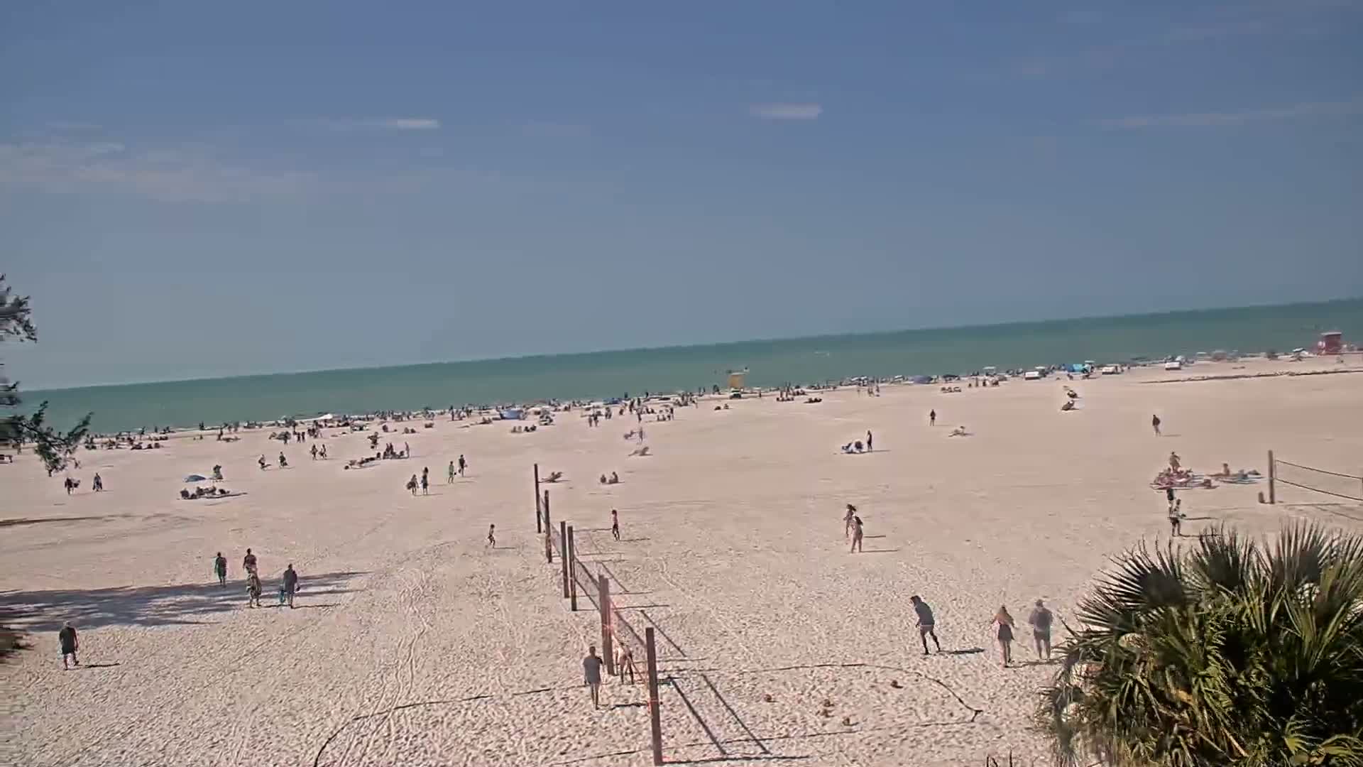 A mostly sunny day on a sandy beach shows many people sunbathing, swimming, and playing volleyball near a calm, light-green ocean.