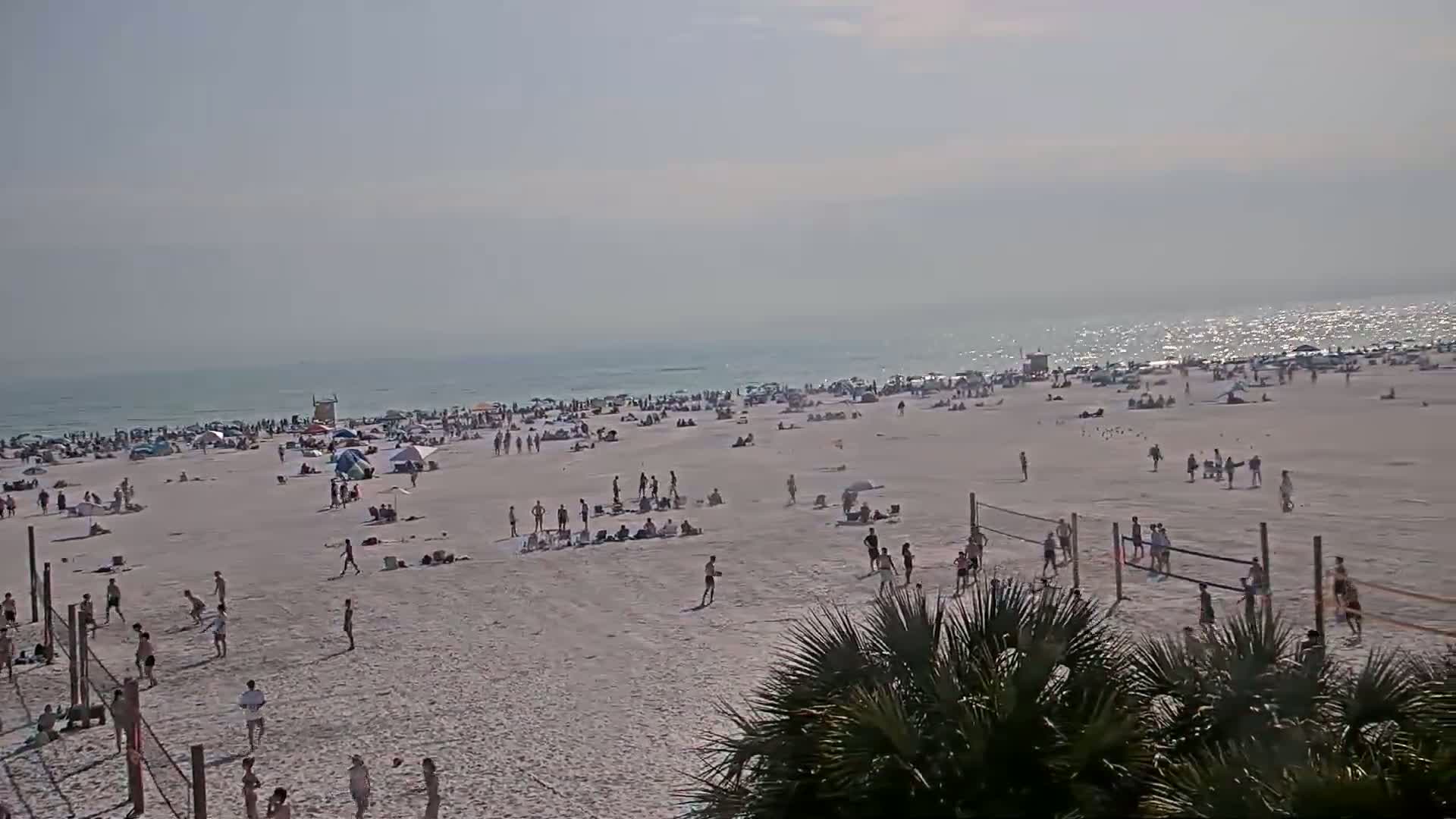 A wide shot shows a crowded beach on a sunny day with calm, sparkling water, people sunbathing, swimming, and playing volleyball.