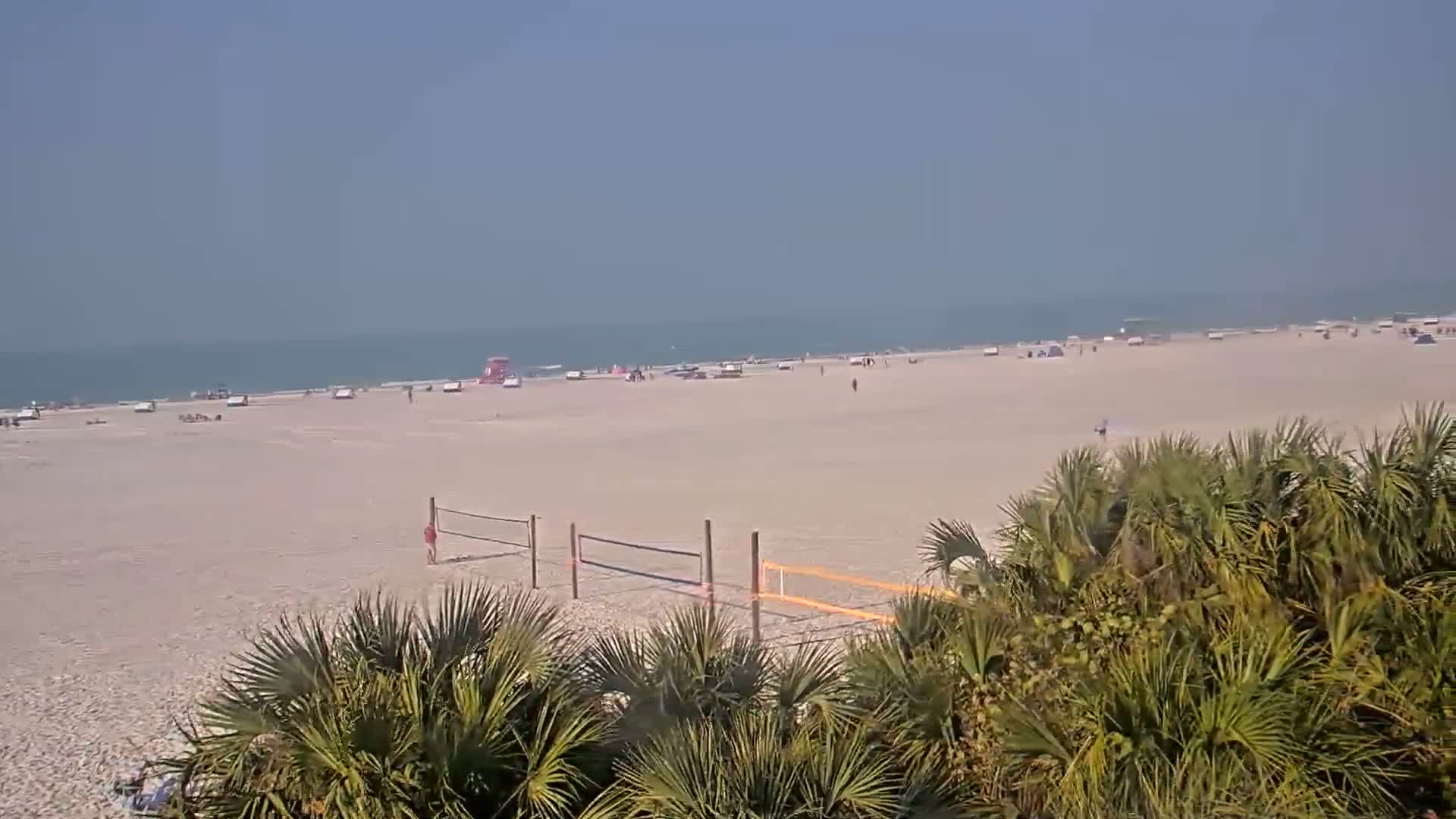 A mostly sunny beach scene shows a wide expanse of light-colored sand with scattered people, a volleyball net, and palm trees in the foreground.