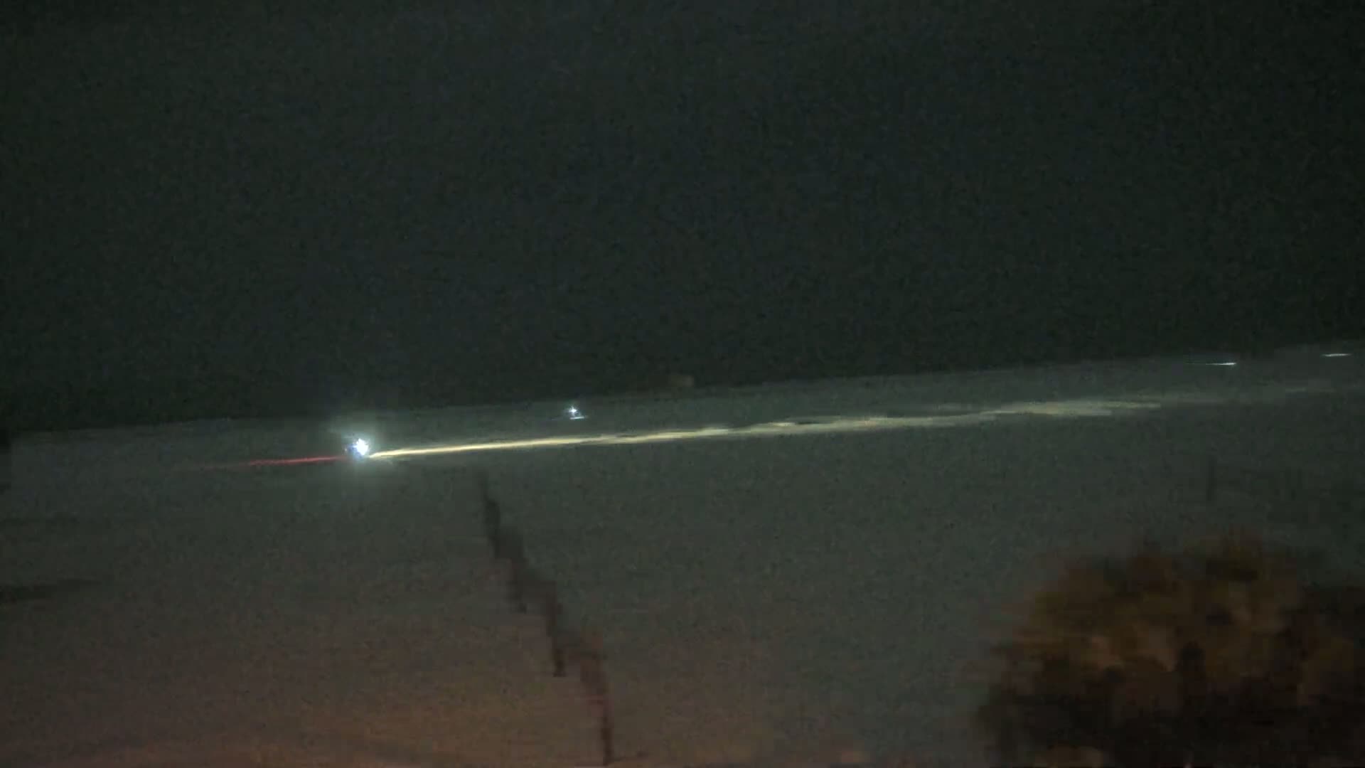 A nighttime view of a snow-covered landscape with a bright light streak across the horizon and faint lights in the distance.