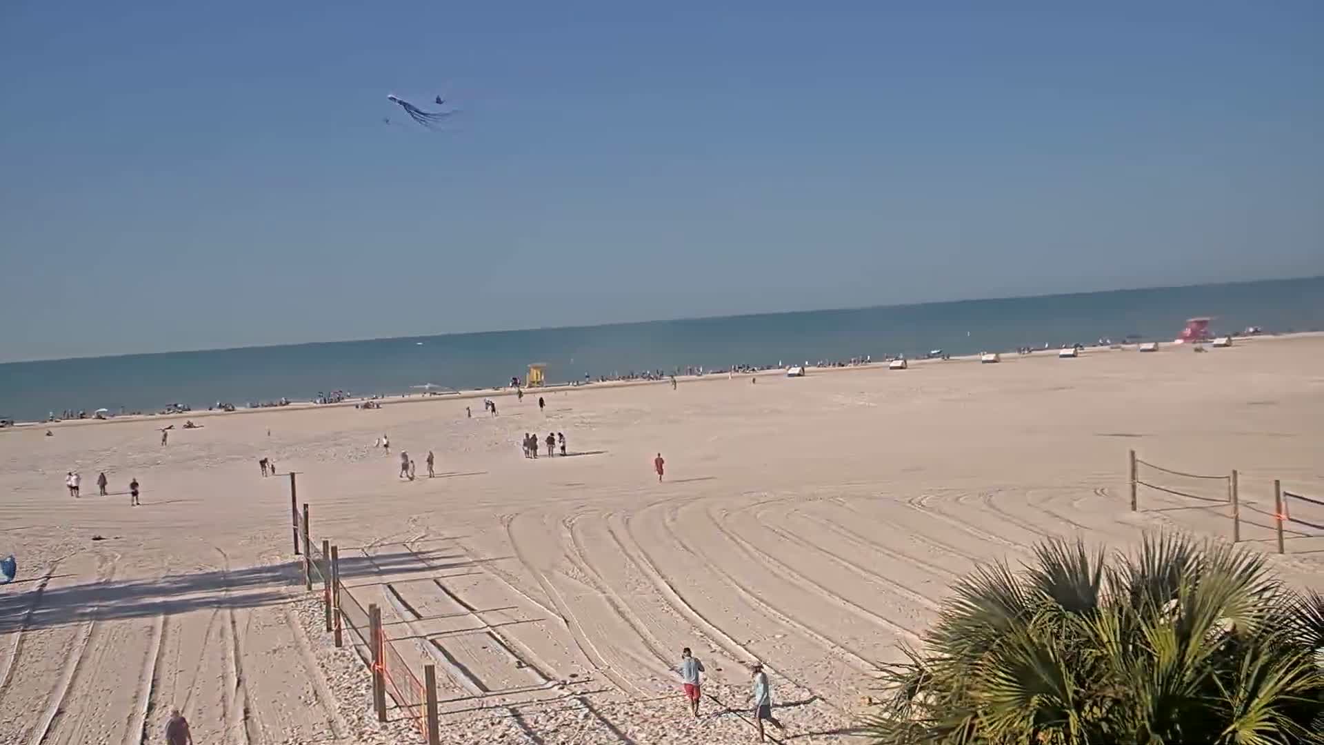A sandy beach with scattered people, volleyball nets, and tire tracks on the sand is visible under a clear blue sky, with a kite flying high above and a calm ocean in the background.