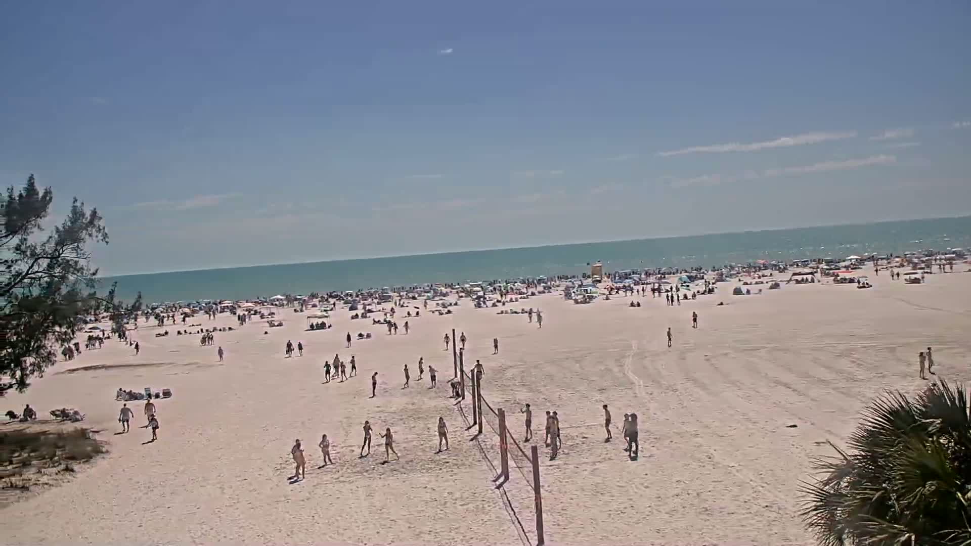 A crowded beach on a sunny day with people playing volleyball and relaxing on the sand near a calm ocean.