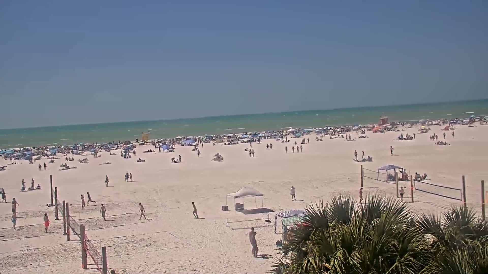 A crowded beach on a sunny day with people sunbathing, playing volleyball, and walking along the shore, with a calm, teal ocean in the background.