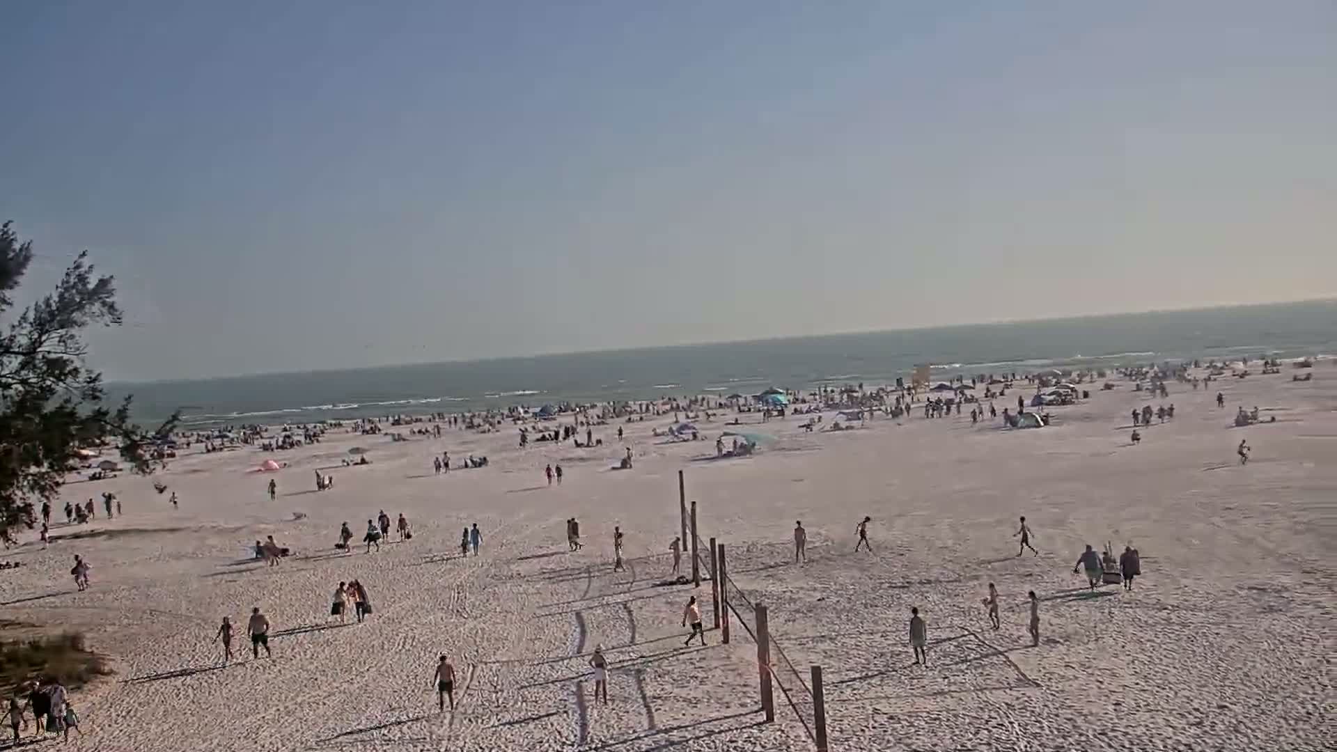 A crowded beach on a sunny day shows many people scattered across the sand, some near the water, others playing volleyball near the center.