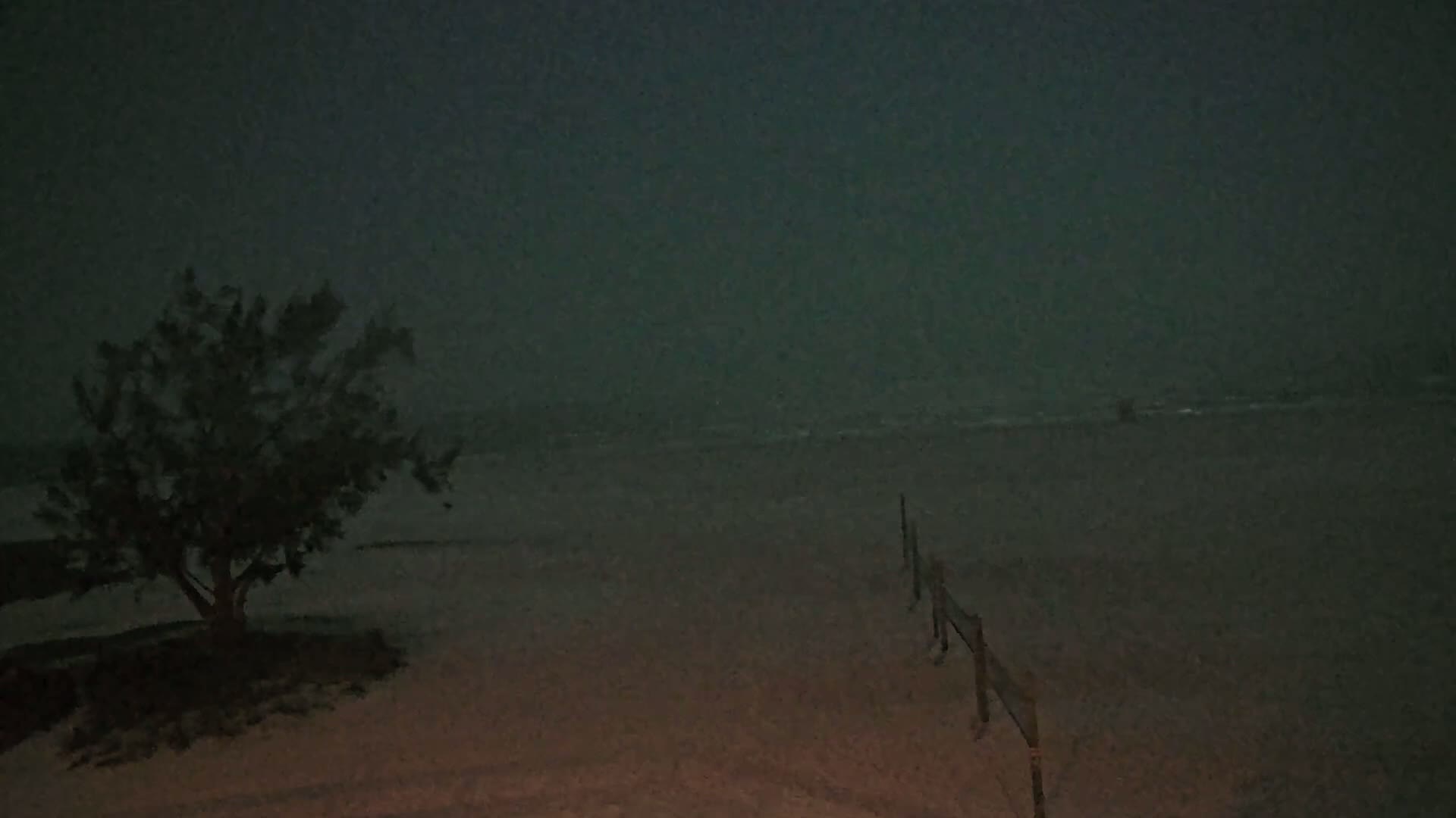 A dark, stormy night scene shows a small tree near a beach with a low wooden fence extending toward a dark ocean.