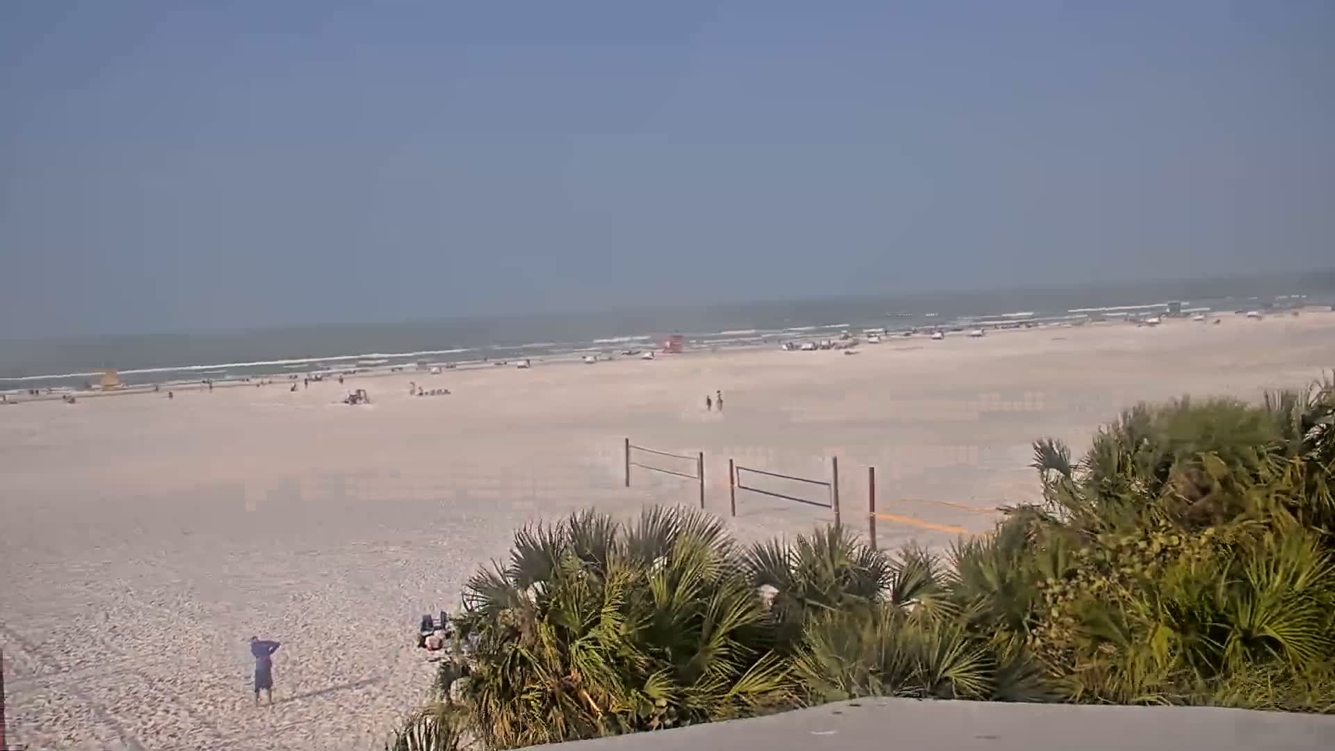 A mostly sunny day at a beach shows a wide expanse of light-brown sand with scattered people, a volleyball net, and palm trees in the foreground.