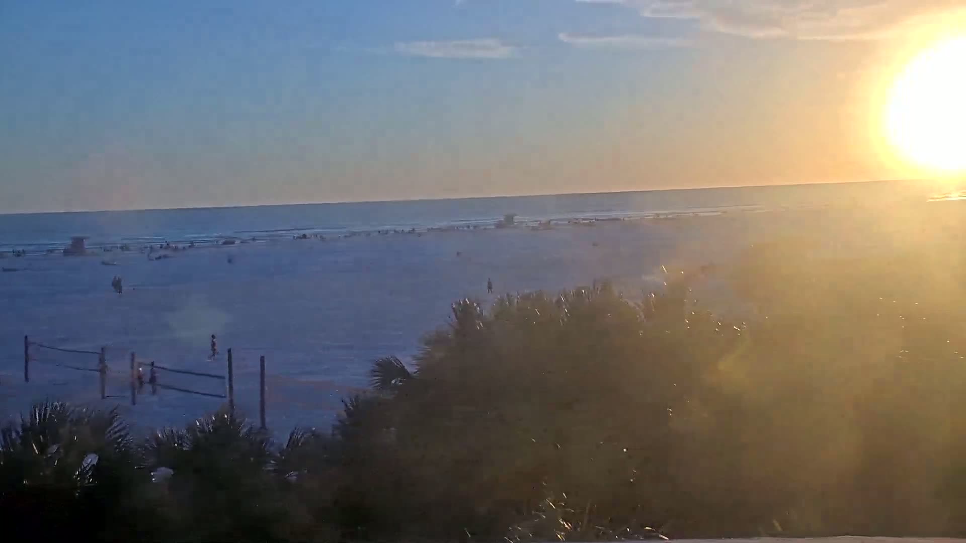 A mostly sunny sunset illuminates a beach scene with people and a volleyball net, viewed from behind shrubbery.