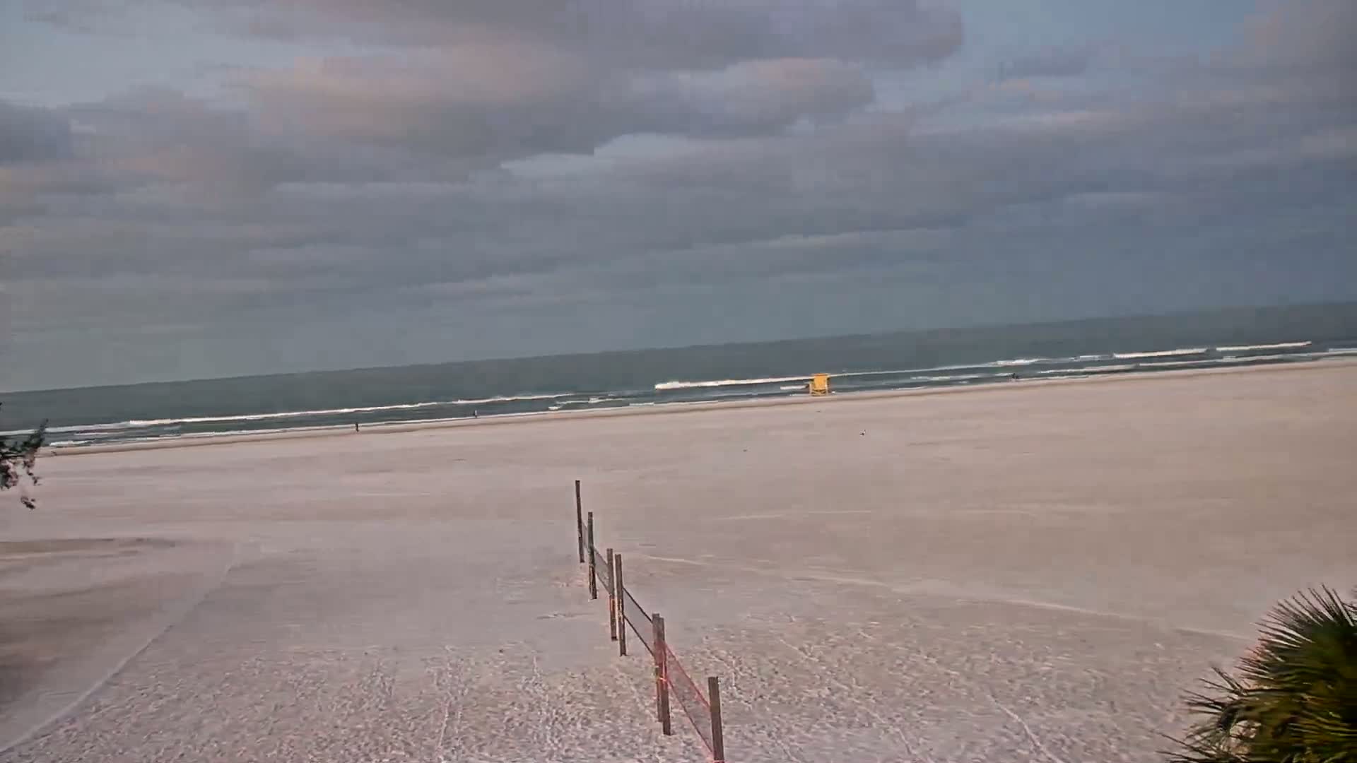 A mostly cloudy day shows a wide expanse of sandy beach leading to a calm ocean with small waves.