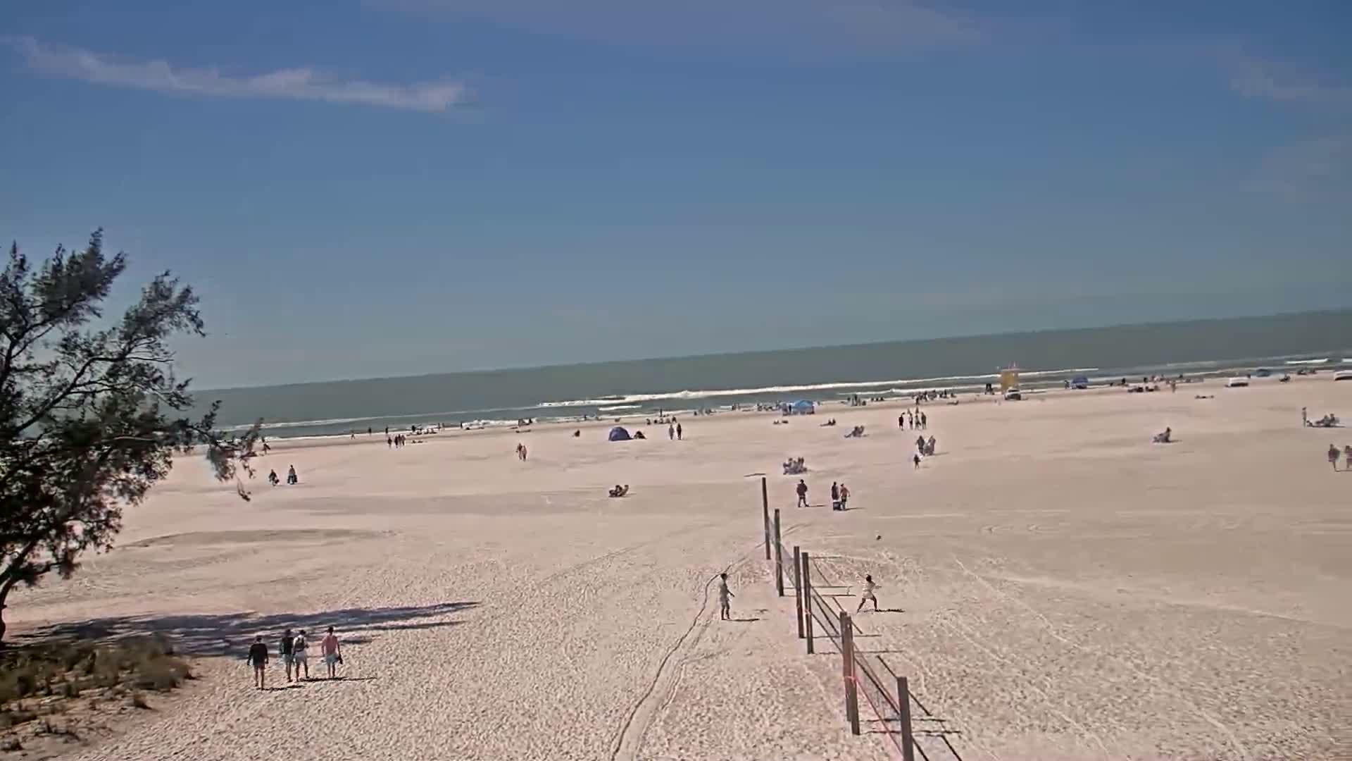 A wide sandy beach with scattered people and a volleyball net stretches toward a calm ocean under a clear blue sky.