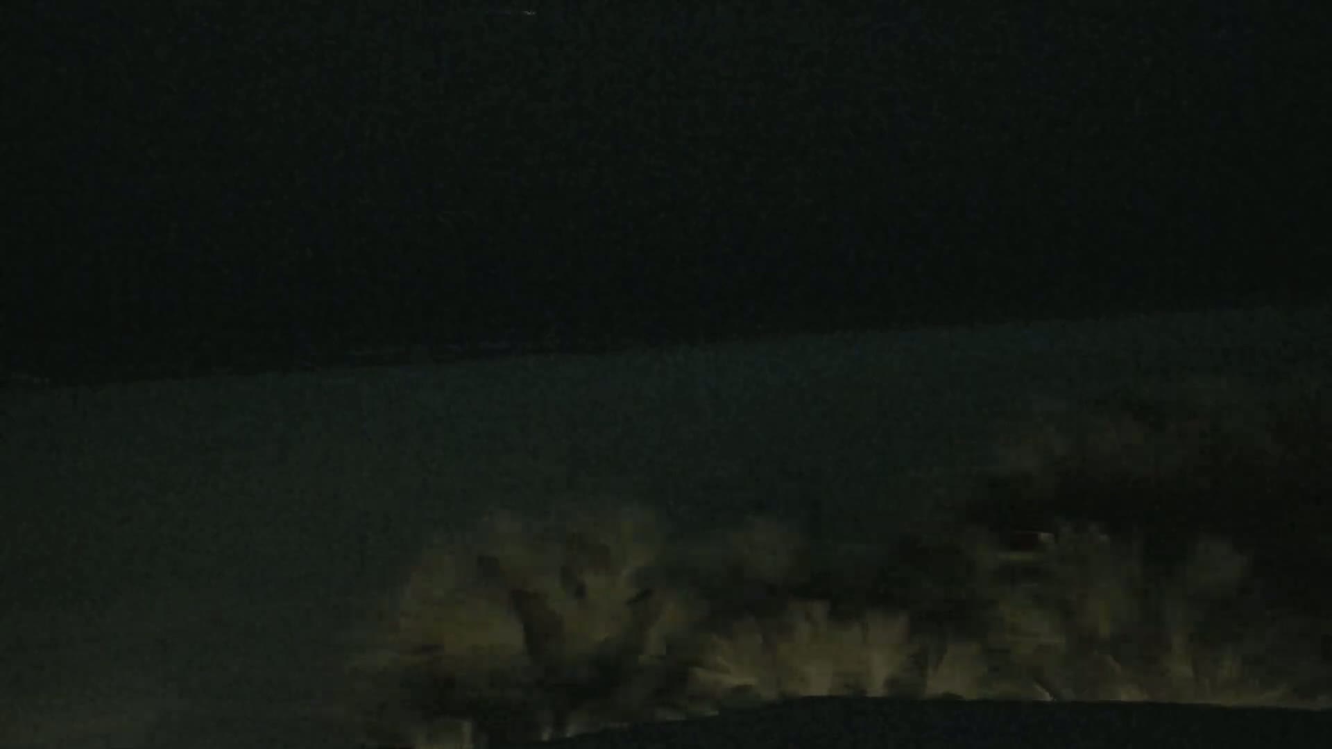 A dark, nighttime scene shows a line of bright, white, possibly foamy waves crashing on a dark shore under a black sky.