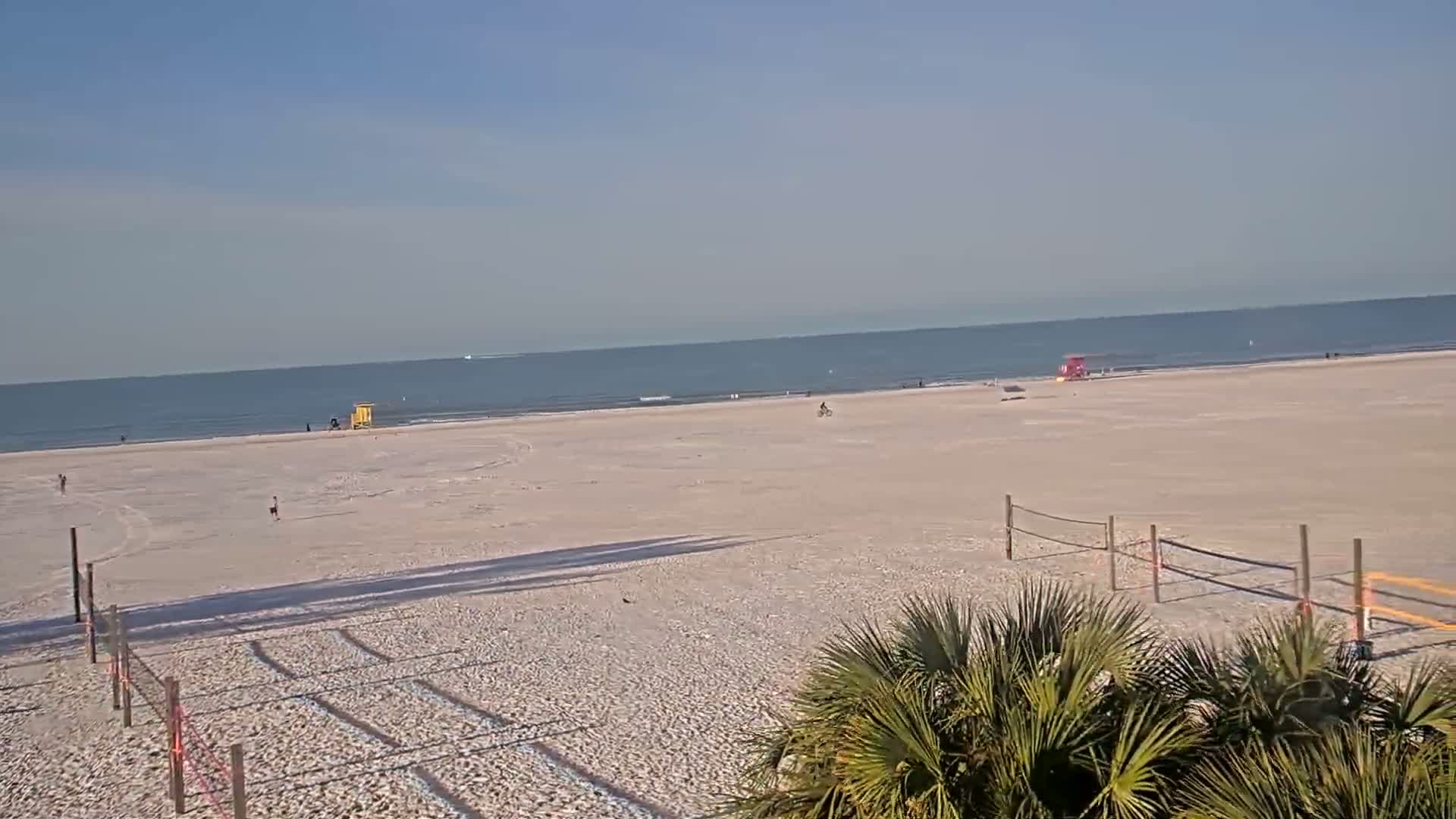A mostly empty, sandy beach stretches to a calm ocean under a clear, bright sky.