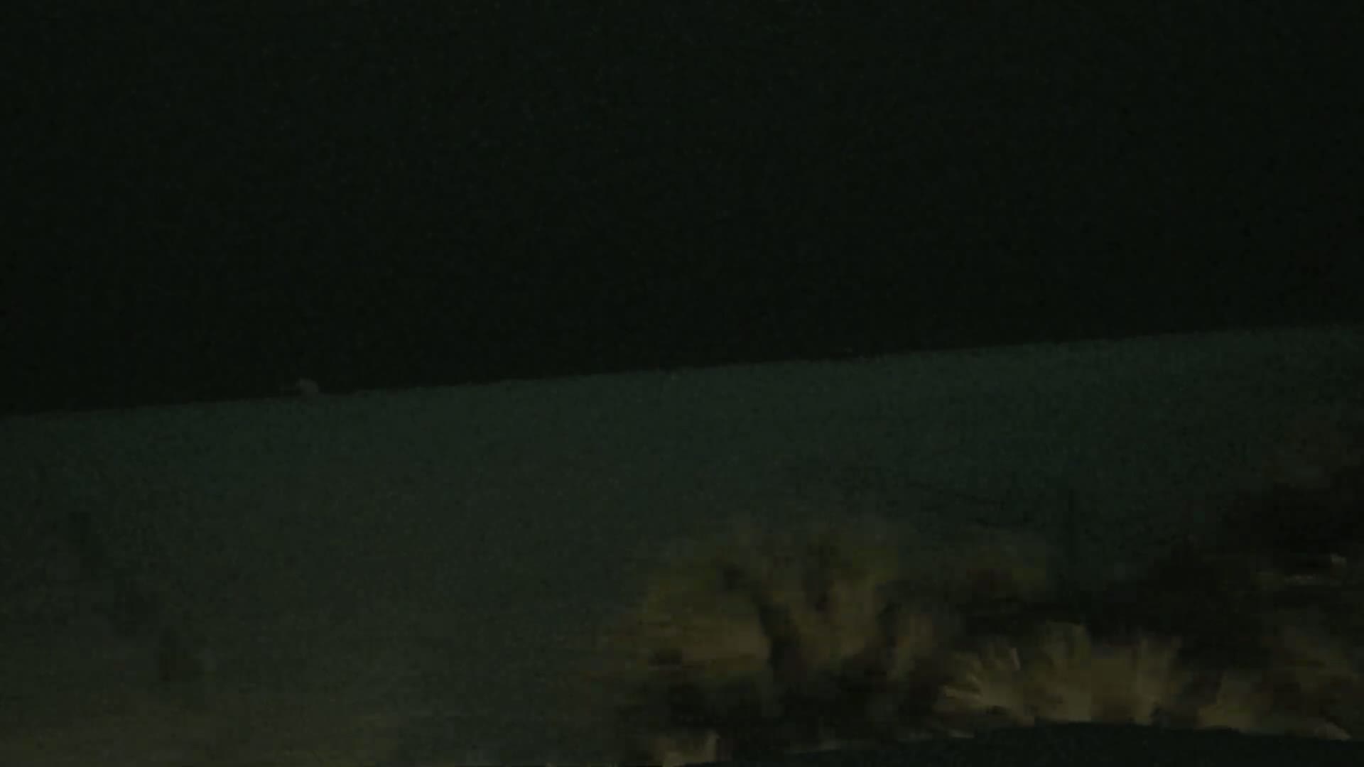 A dark, nighttime scene shows a body of water in the distance with vegetation in the foreground under a dark, possibly stormy sky.