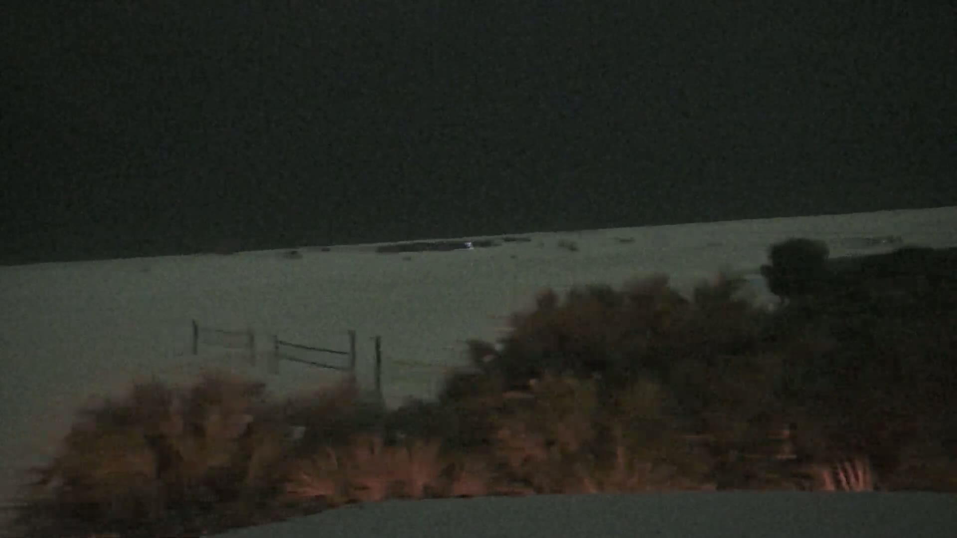 A nighttime view of a snow-covered beach seen from behind a line of shrubs and a fence.