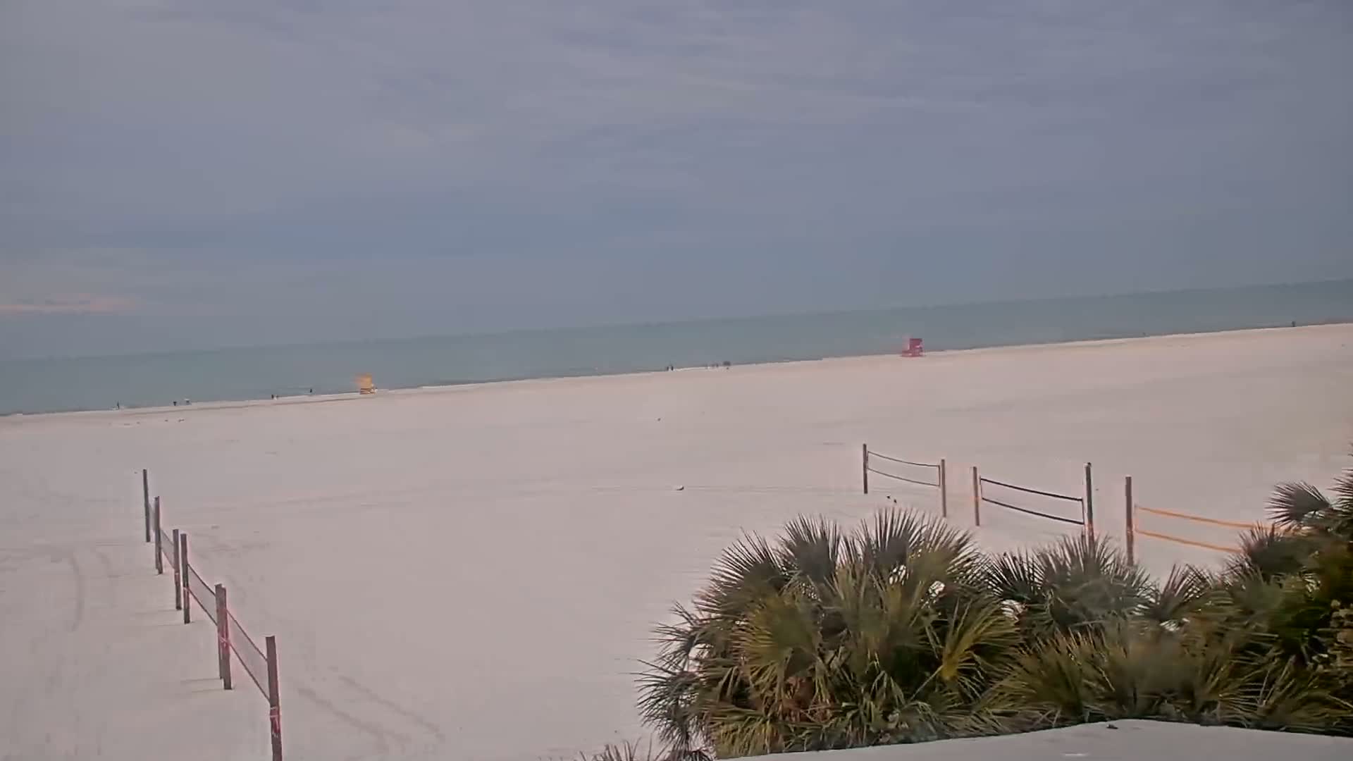 A mostly empty, white sandy beach stretches to a calm, light-grey ocean under an overcast sky, with volleyball nets and palm trees in the foreground.