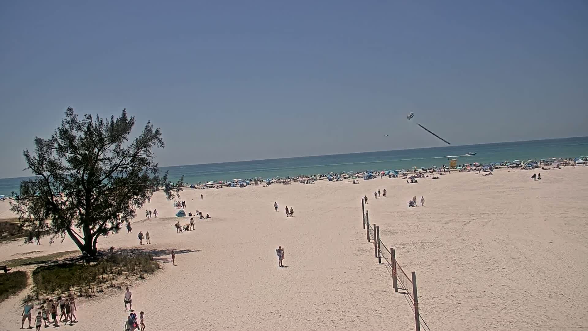 A sandy beach with scattered people, umbrellas, and a volleyball net under a clear blue sky, with a dark object trailing in the sky above the ocean.