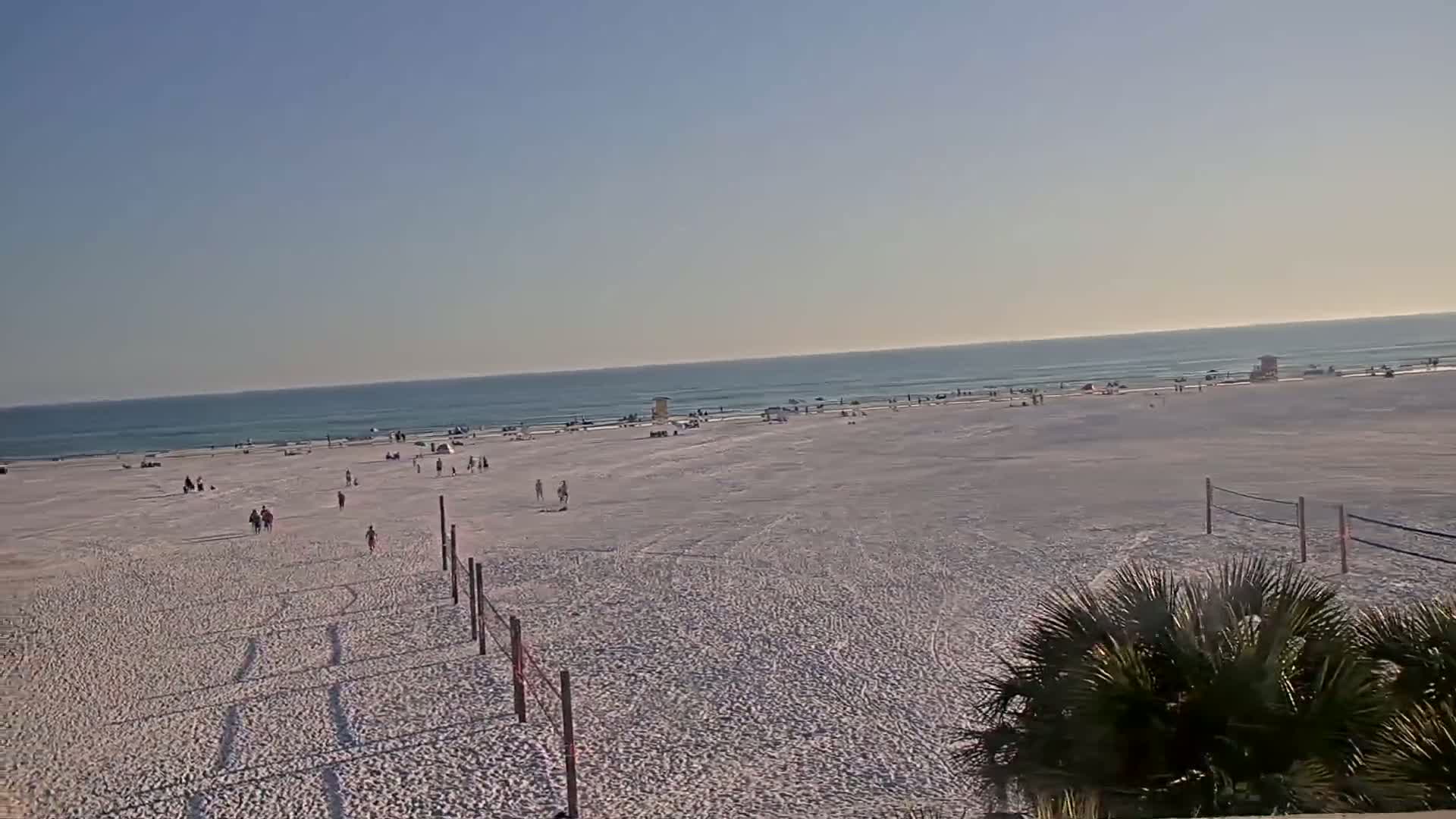 A wide sandy beach with scattered people and volleyball nets stretches to a calm ocean under a clear blue sky.