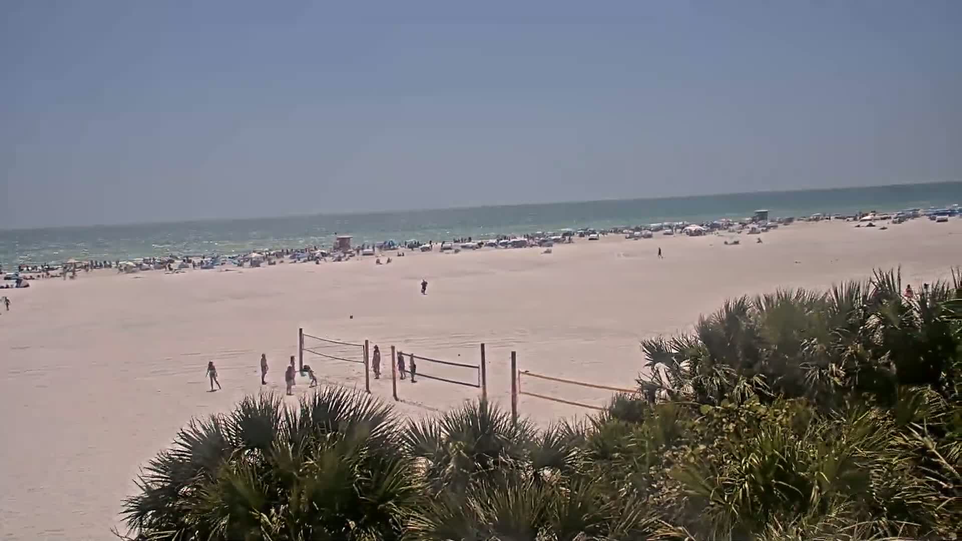 A mostly sunny beach scene shows a wide expanse of white sand with scattered people, a volleyball net, and palm trees in the foreground, with a calm ocean in the background.