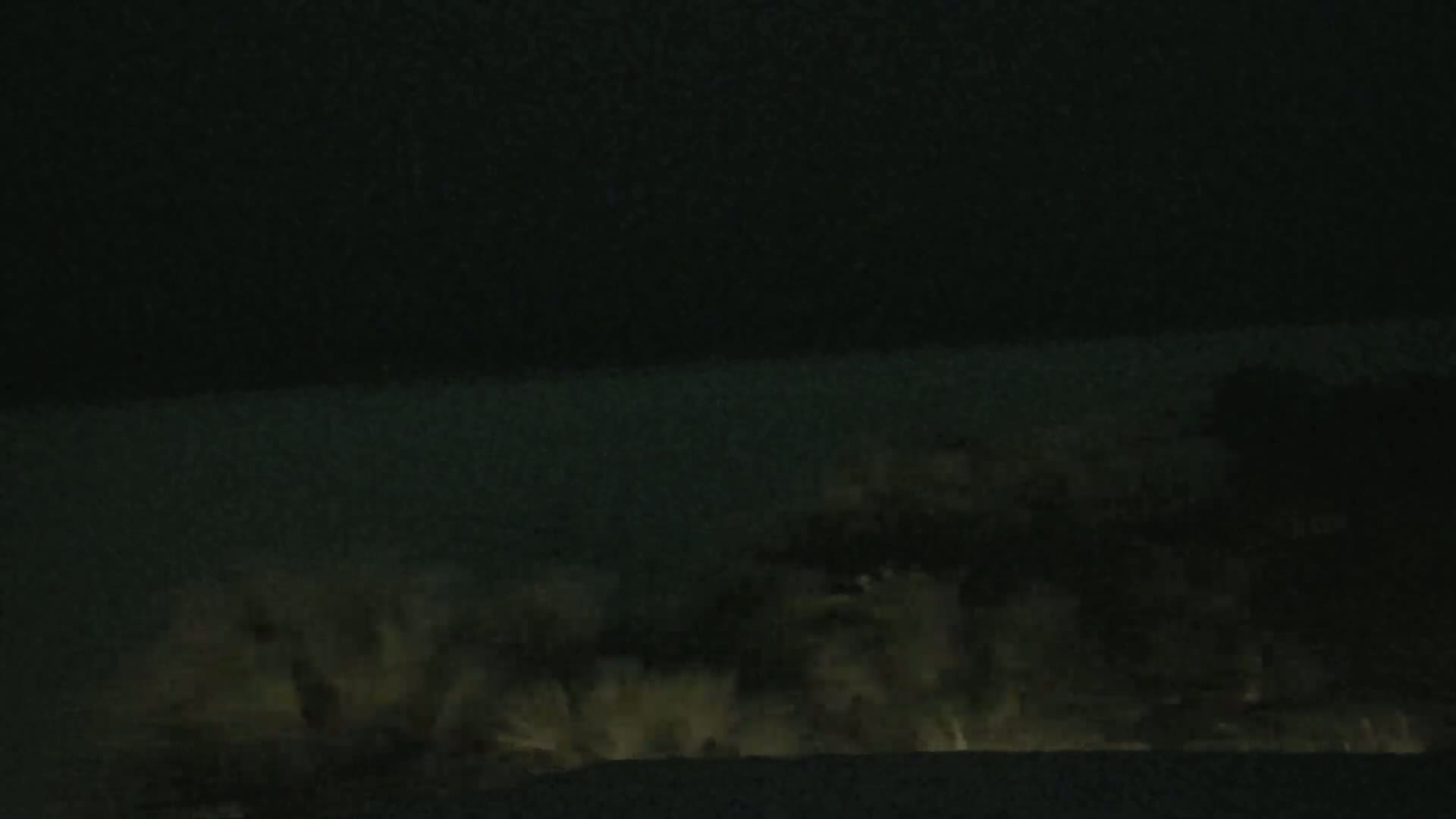A dark, nighttime scene shows a body of water with waves crashing against a shoreline, lit faintly from below.