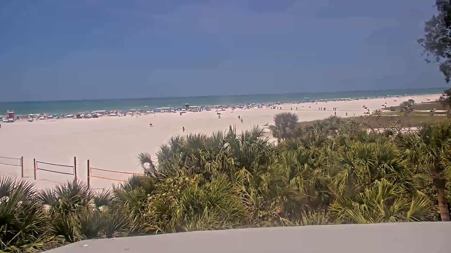 A sunny beach scene shows a wide expanse of sand with many people, some beach umbrellas, and a volleyball net, all overlooked by low-lying vegetation.
