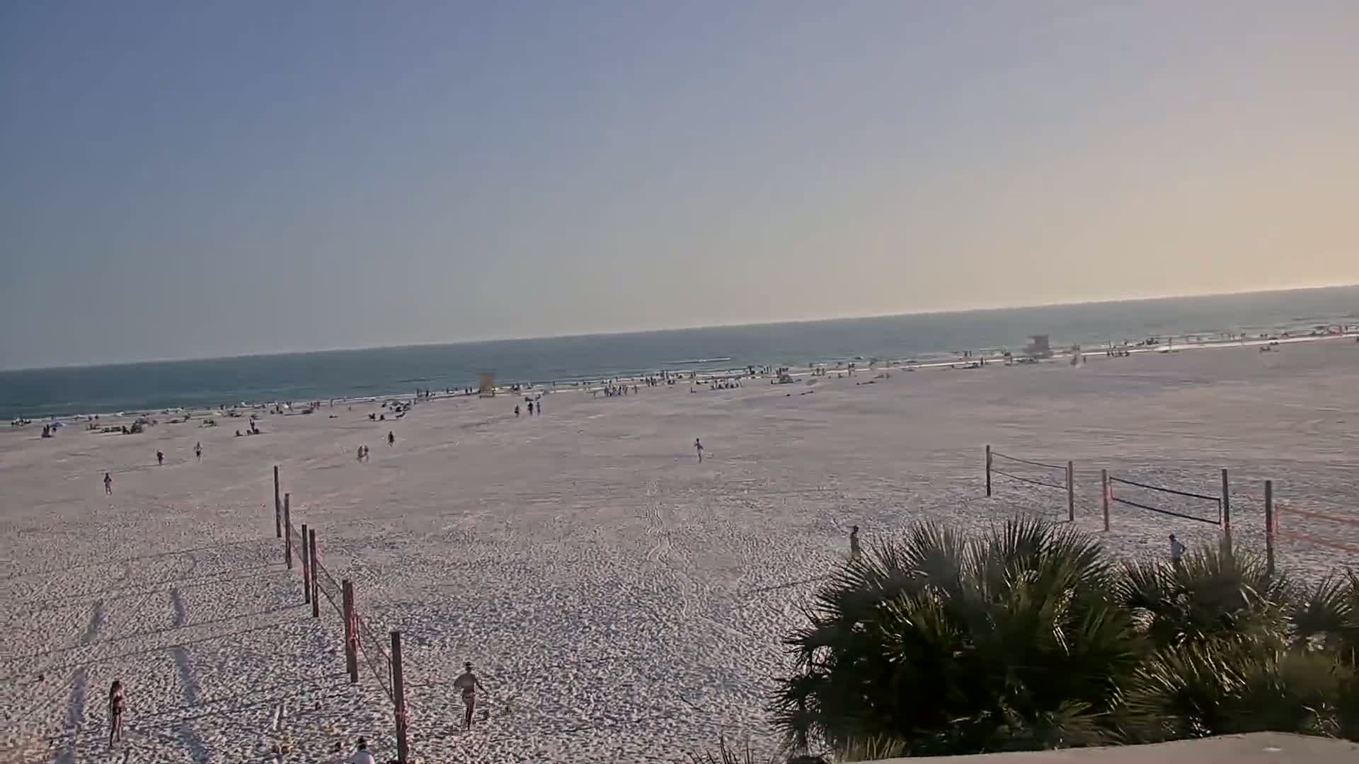A mostly sunny beach scene shows a wide expanse of light-colored sand with scattered people, volleyball nets, and a calm ocean in the background.