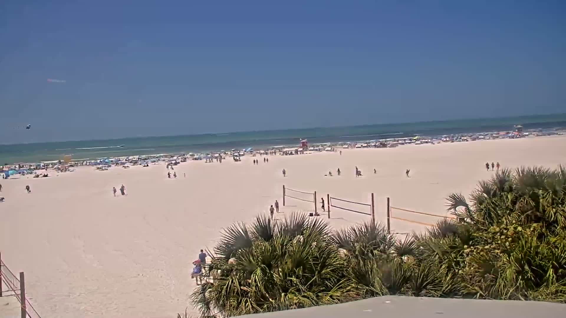 A wide sandy beach with many people and a volleyball net is shown, under a clear blue sky; the ocean is calm with small waves.