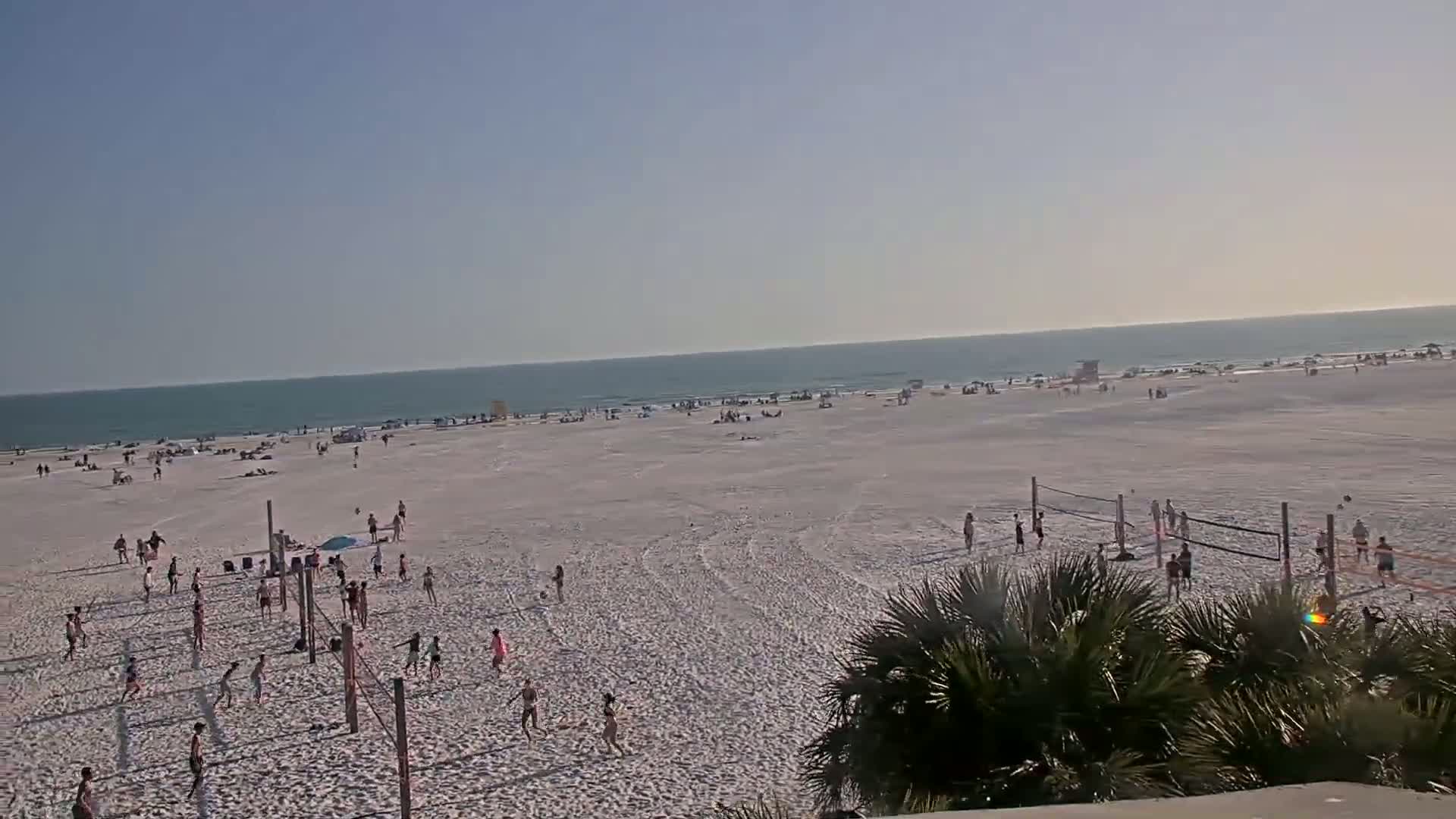 A wide shot of a sandy beach with many people playing volleyball and relaxing on a sunny day, the ocean visible in the background.