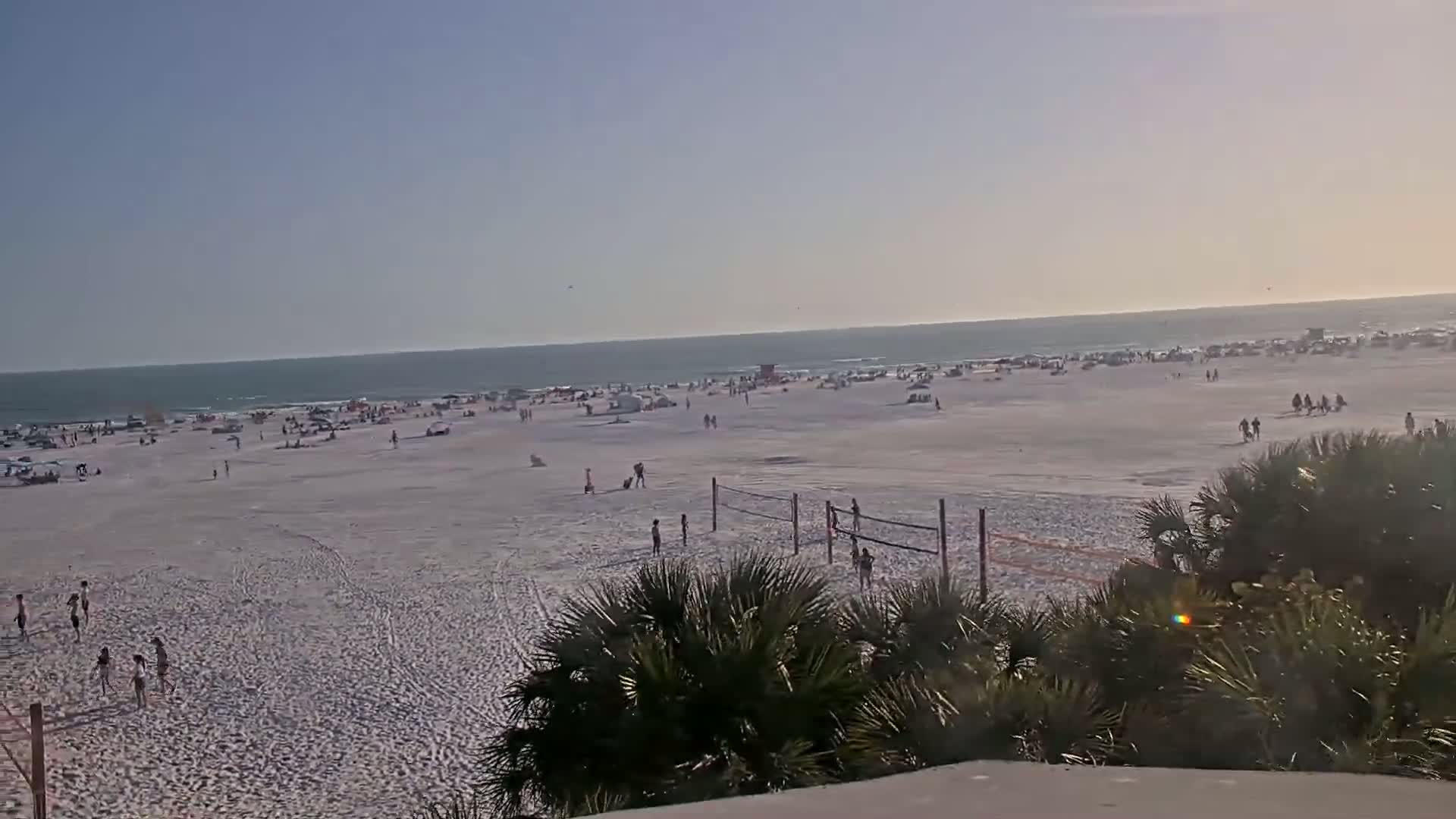 A wide shot of a crowded beach on a sunny day shows people relaxing, playing volleyball, and walking near the ocean.
