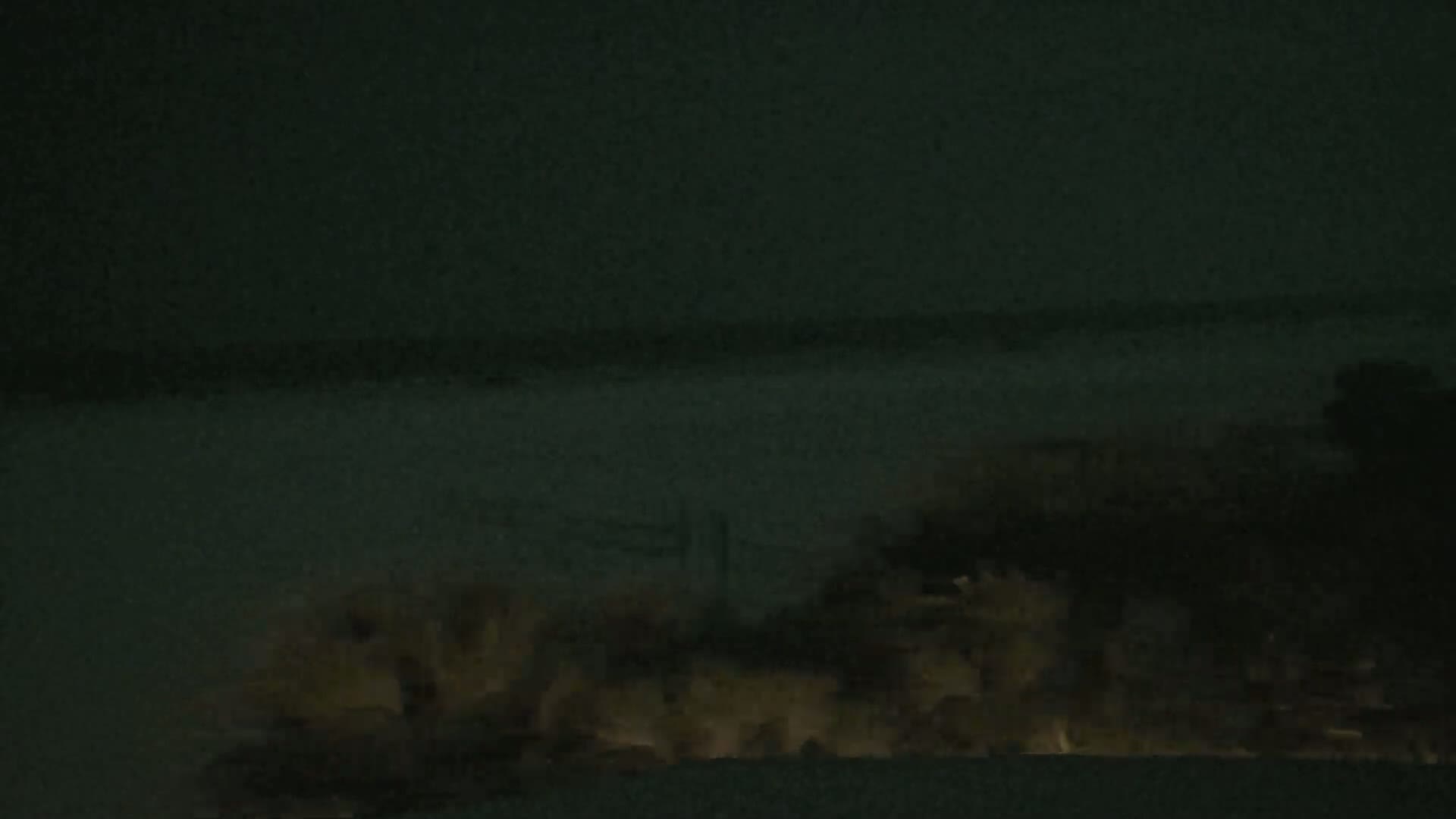 A dark, nighttime scene shows dimly lit, blurry objects in the lower half of the frame, possibly buildings or vegetation, against a dark, featureless background.