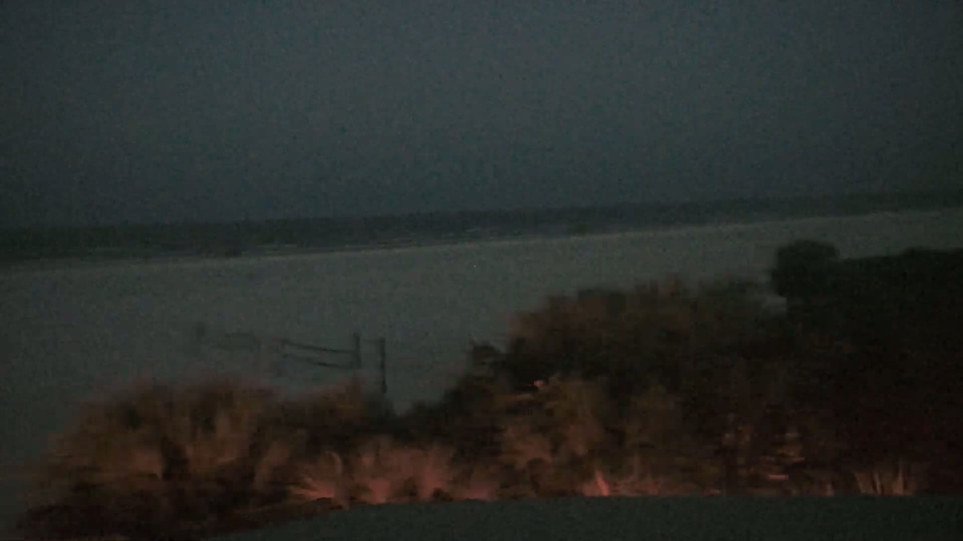 A dark, overcast sky looms over a beach scene at night, where windblown grasses glow faintly in the low light, with a calm, dark ocean in the distance.