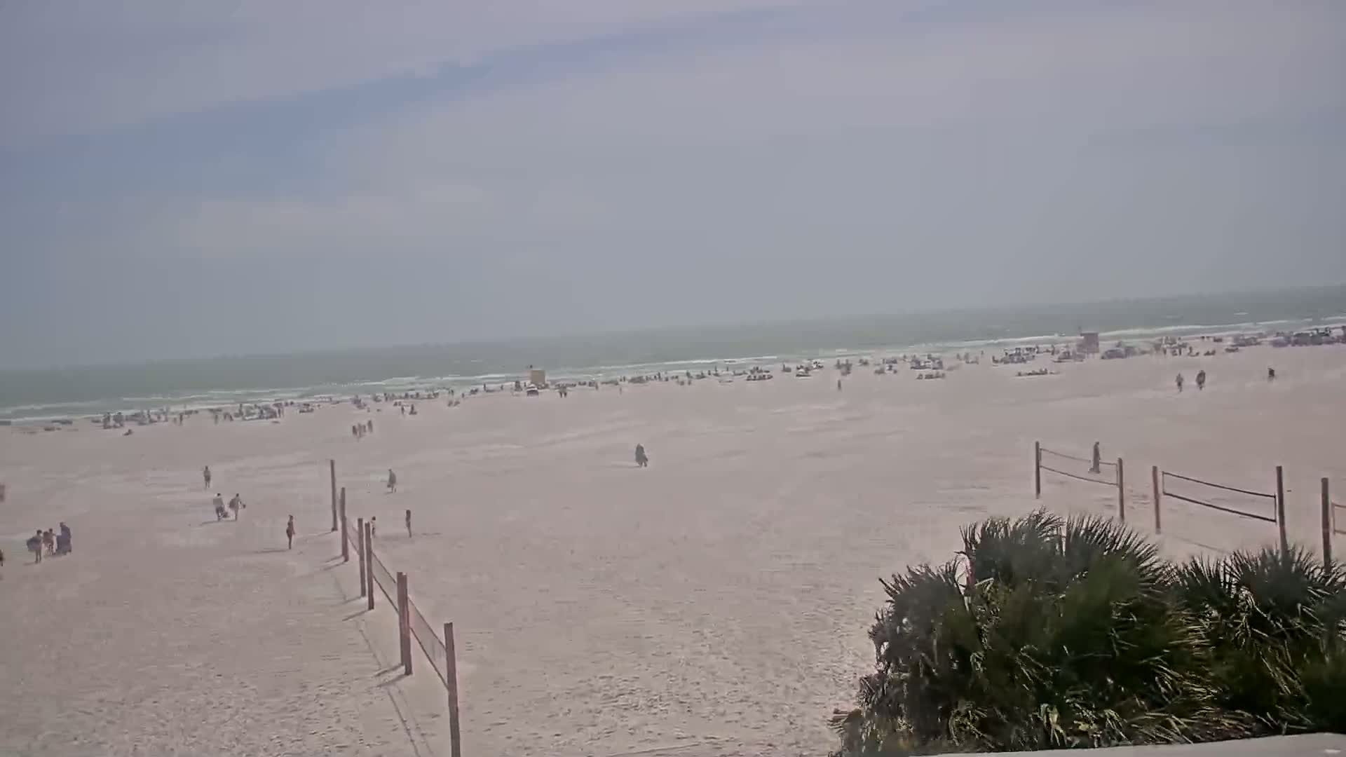 A wide shot of a beach with many people scattered across the sand under a hazy sky and a calm ocean.