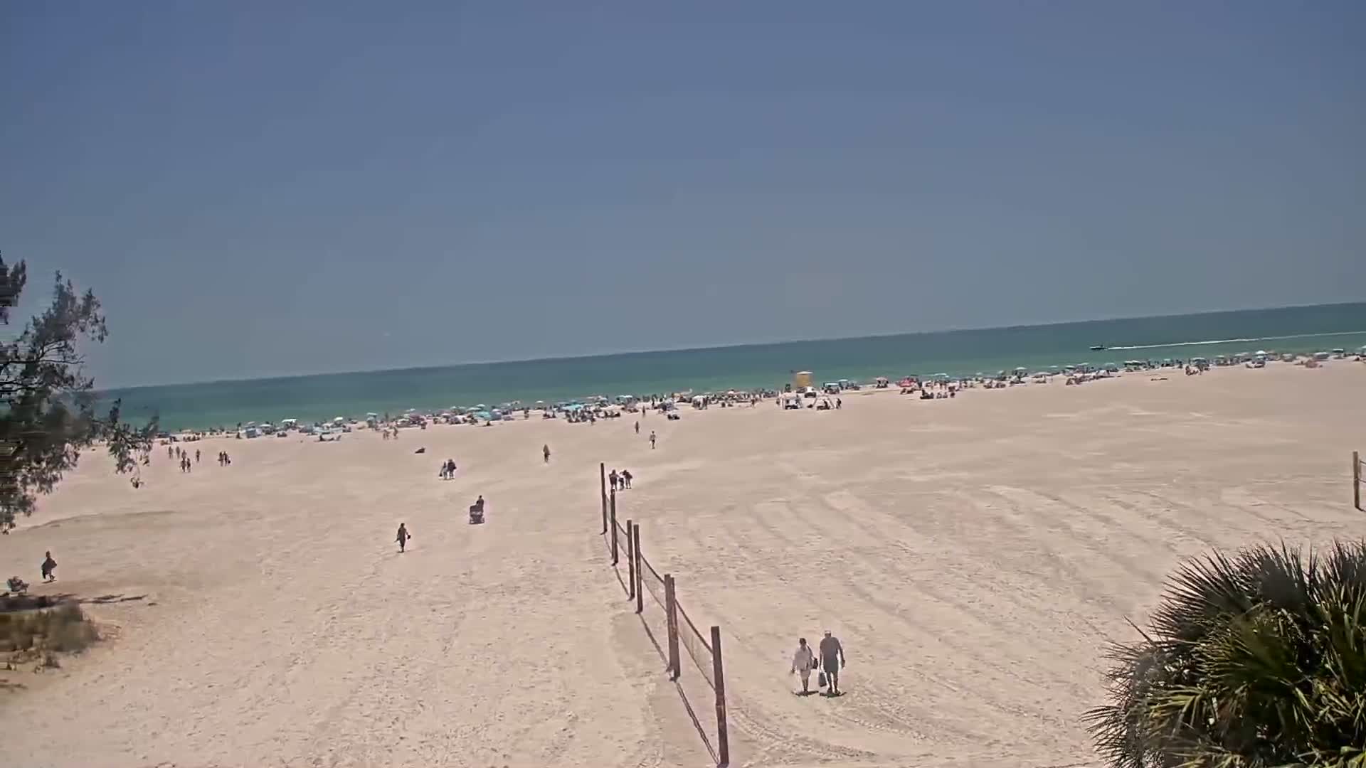 A mostly sunny day shows a wide expanse of sandy beach with scattered people and beach umbrellas, leading to a calm, light-blue ocean.