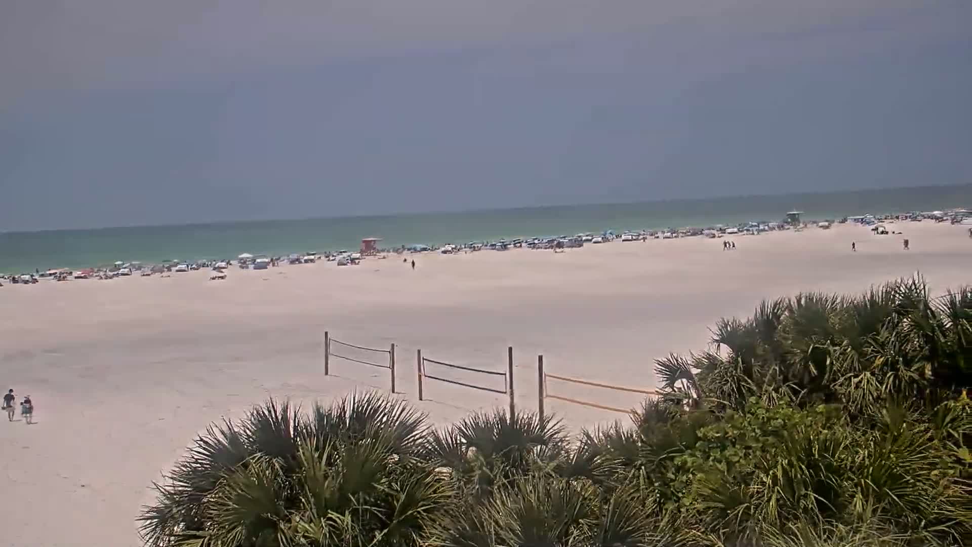 A mostly sunny beach scene shows a wide expanse of light-sand beach with numerous people and parked cars near the water, a volleyball net, and some palm trees in the foreground.
