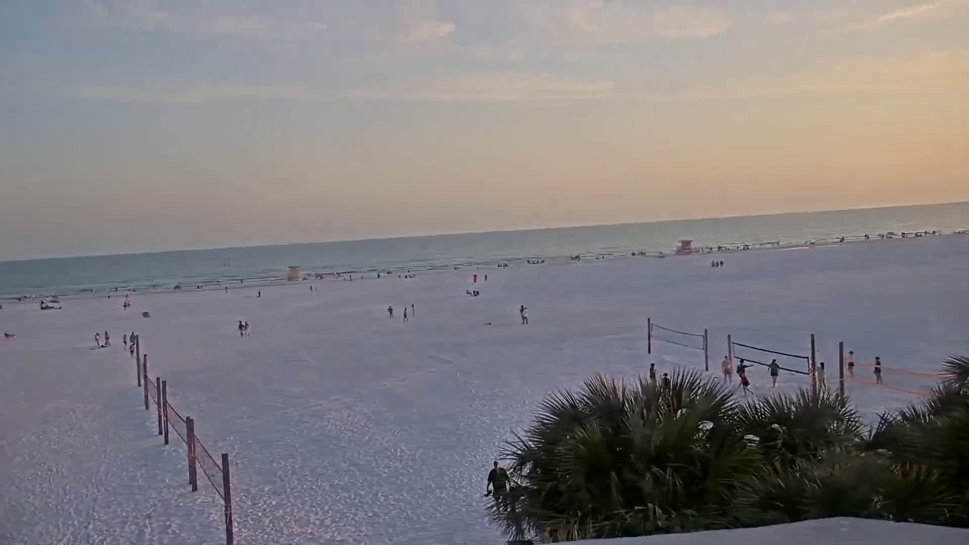 A wide, sandy beach with scattered people, volleyball nets, and palm trees in the foreground, under a pale sunset sky.