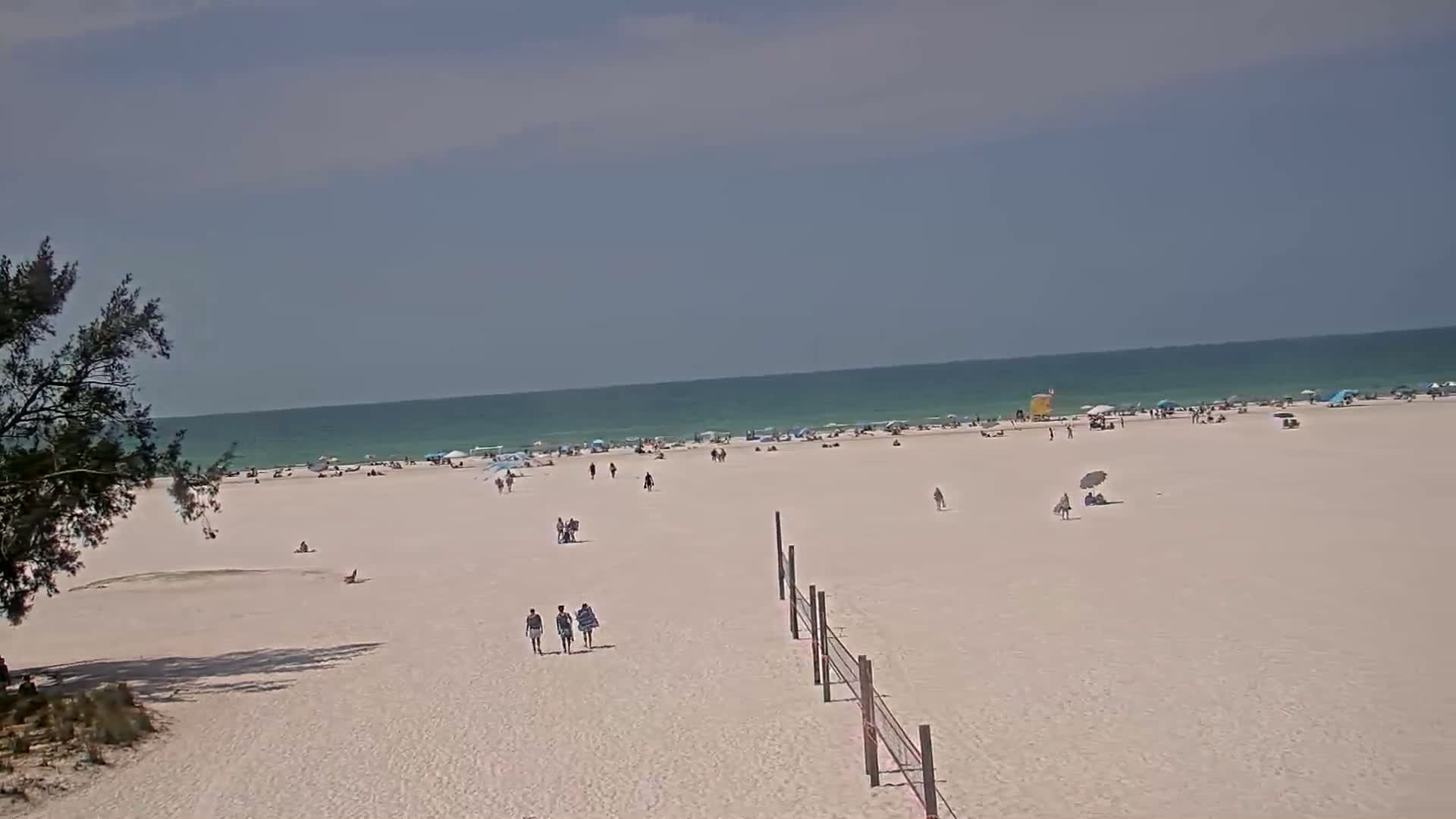 A mostly sunny day at a beach shows a wide expanse of white sand with scattered people, umbrellas, and a volleyball net, and a calm, light-green ocean in the background.