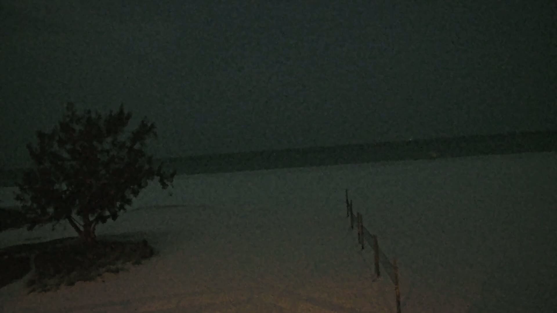 A dark, nighttime scene shows a small tree near a snow-covered beach with a short fence extending toward a dark ocean under a clouded sky.