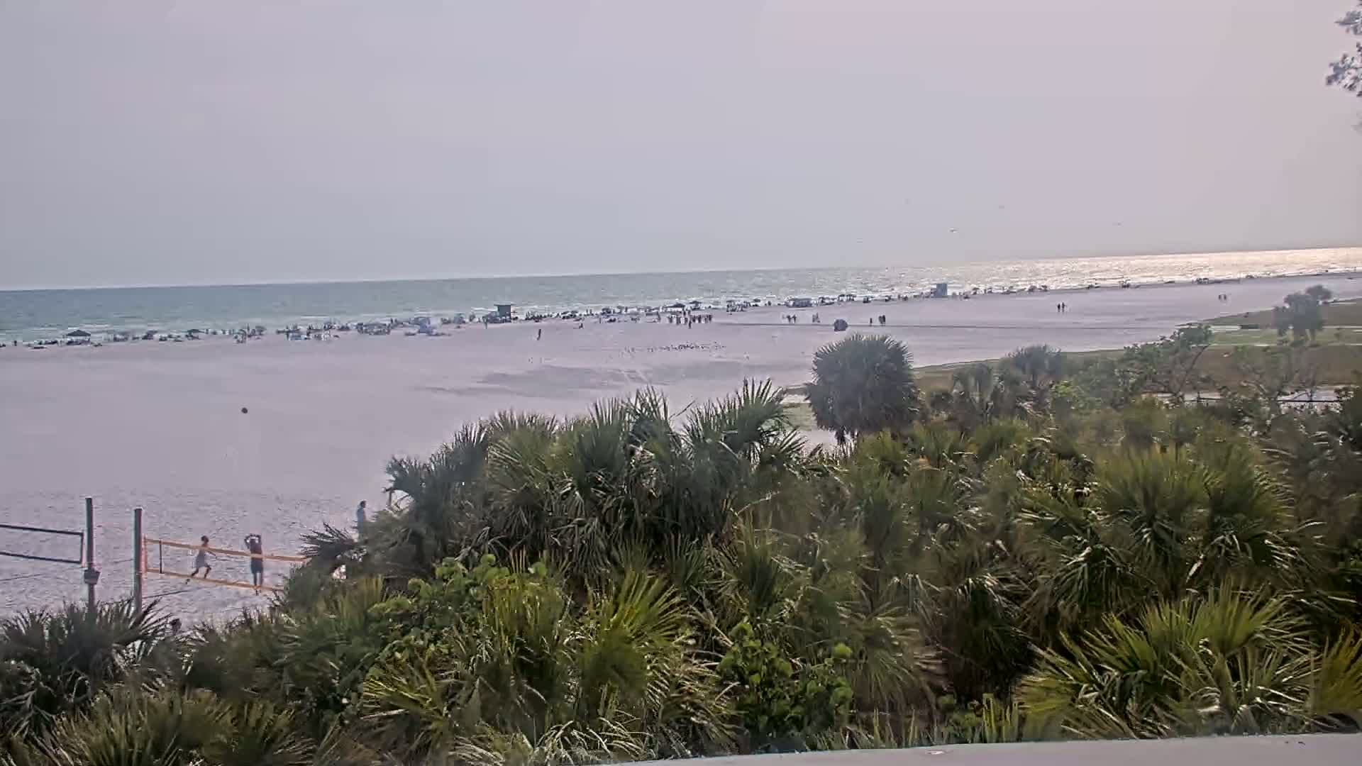 A mostly sunny beach scene shows people scattered on the sand and in the water, with a volleyball net in the foreground and lush green vegetation between the viewer and the beach.