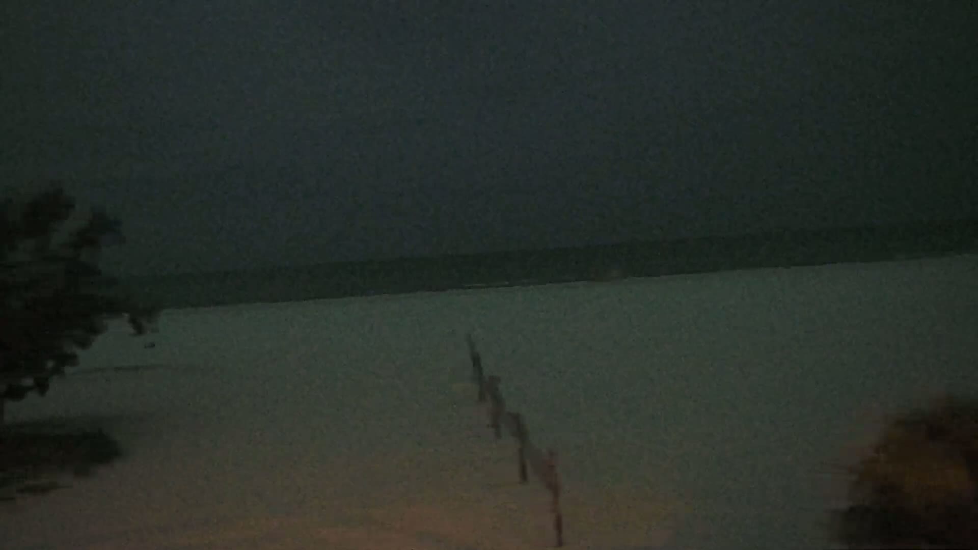 A dark, nighttime beach scene shows a calm ocean under a cloudy sky, with a line of posts leading from the water's edge towards vegetation.