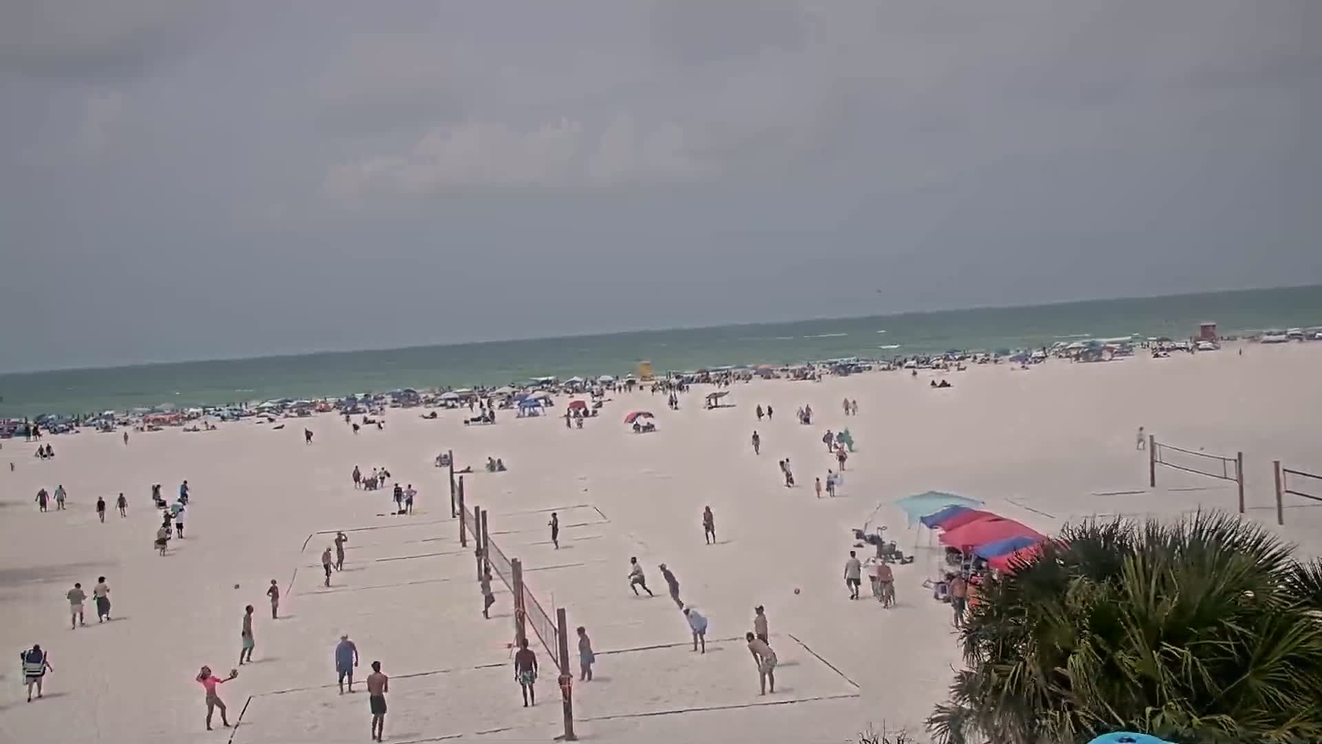 A wide, sandy beach with many people playing volleyball and relaxing under umbrellas, in front of a calm, light-green ocean under an overcast sky.
