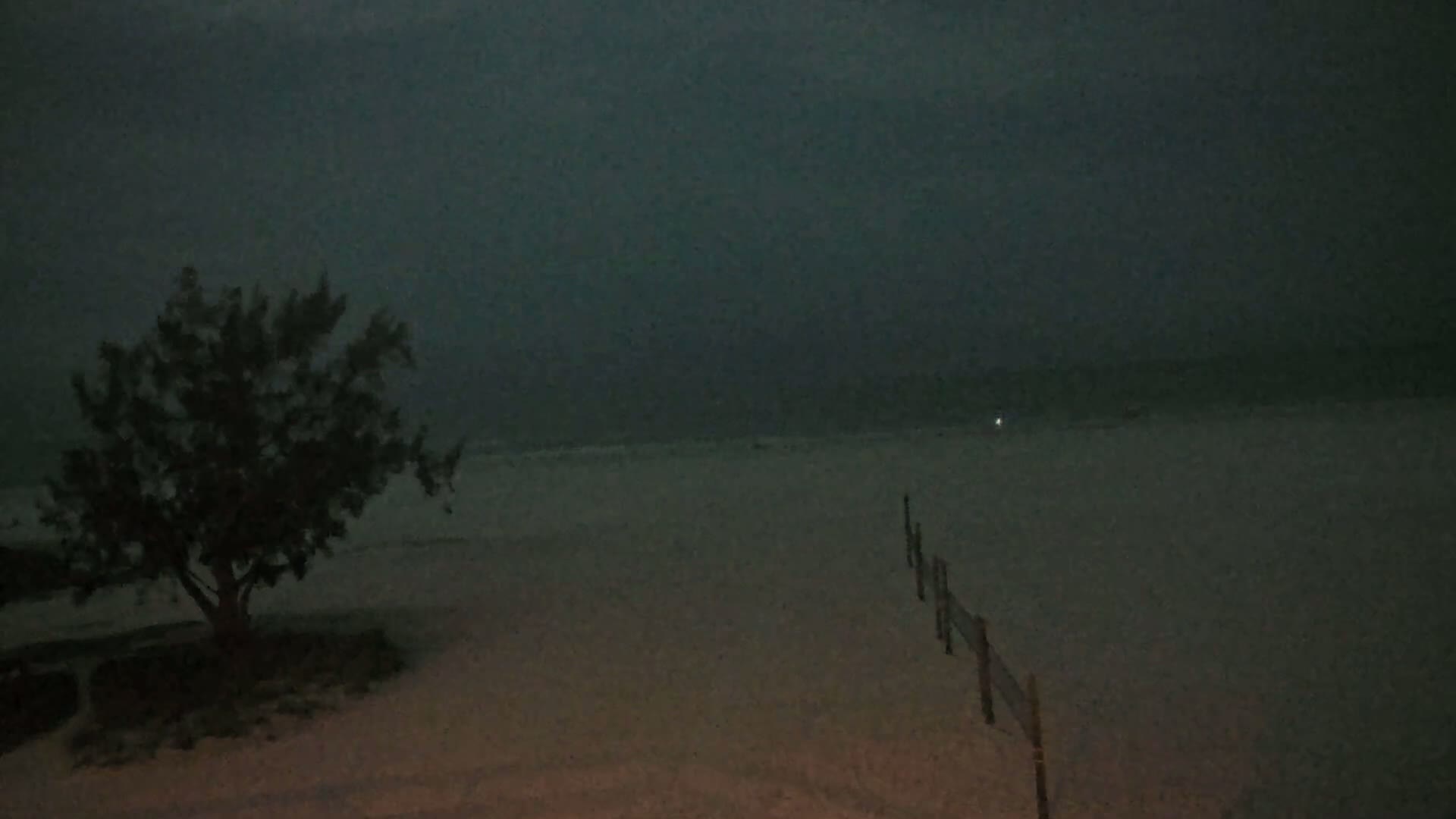 A dark, stormy night scene shows a small tree near a beach with a wooden fence extending toward a dimly lit ocean under a gray sky.