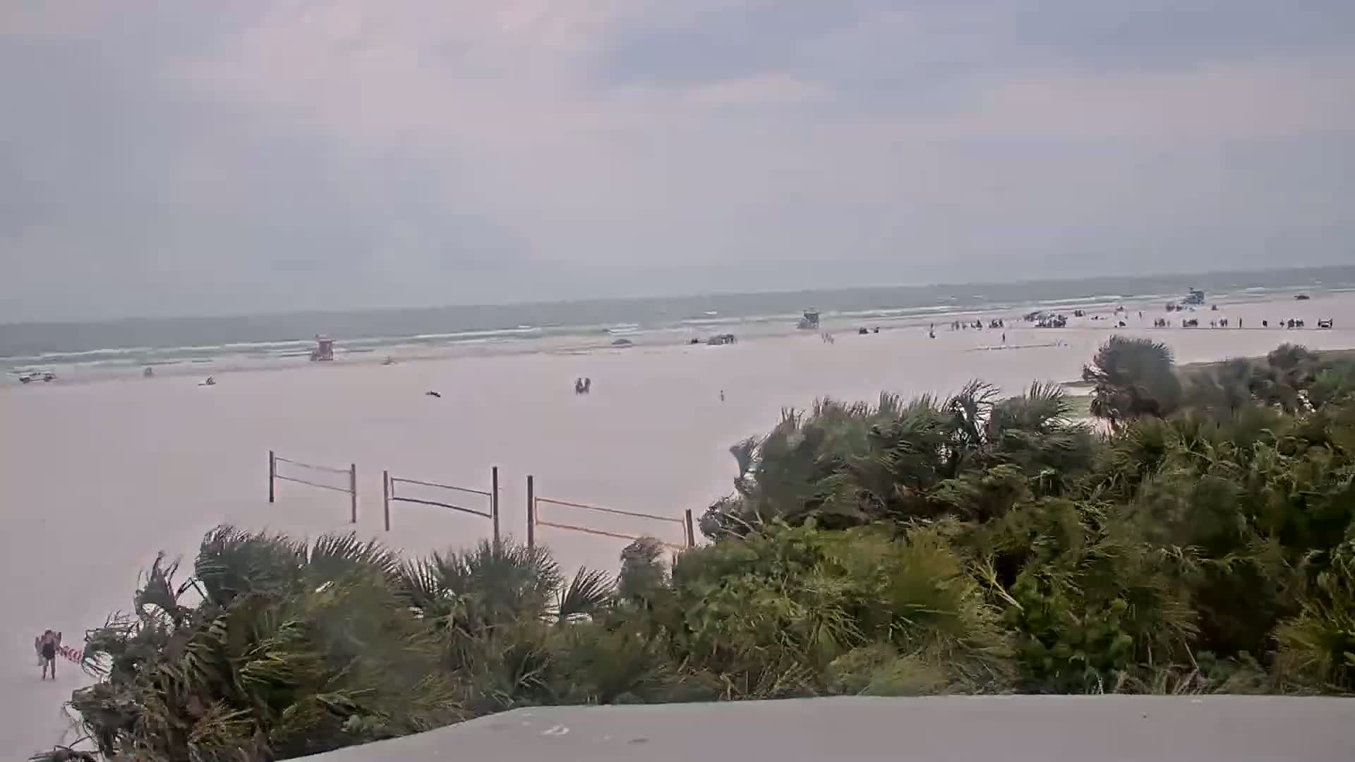 A mostly empty sandy beach with a few people and volleyball nets is seen from behind a line of windswept vegetation, under an overcast sky.