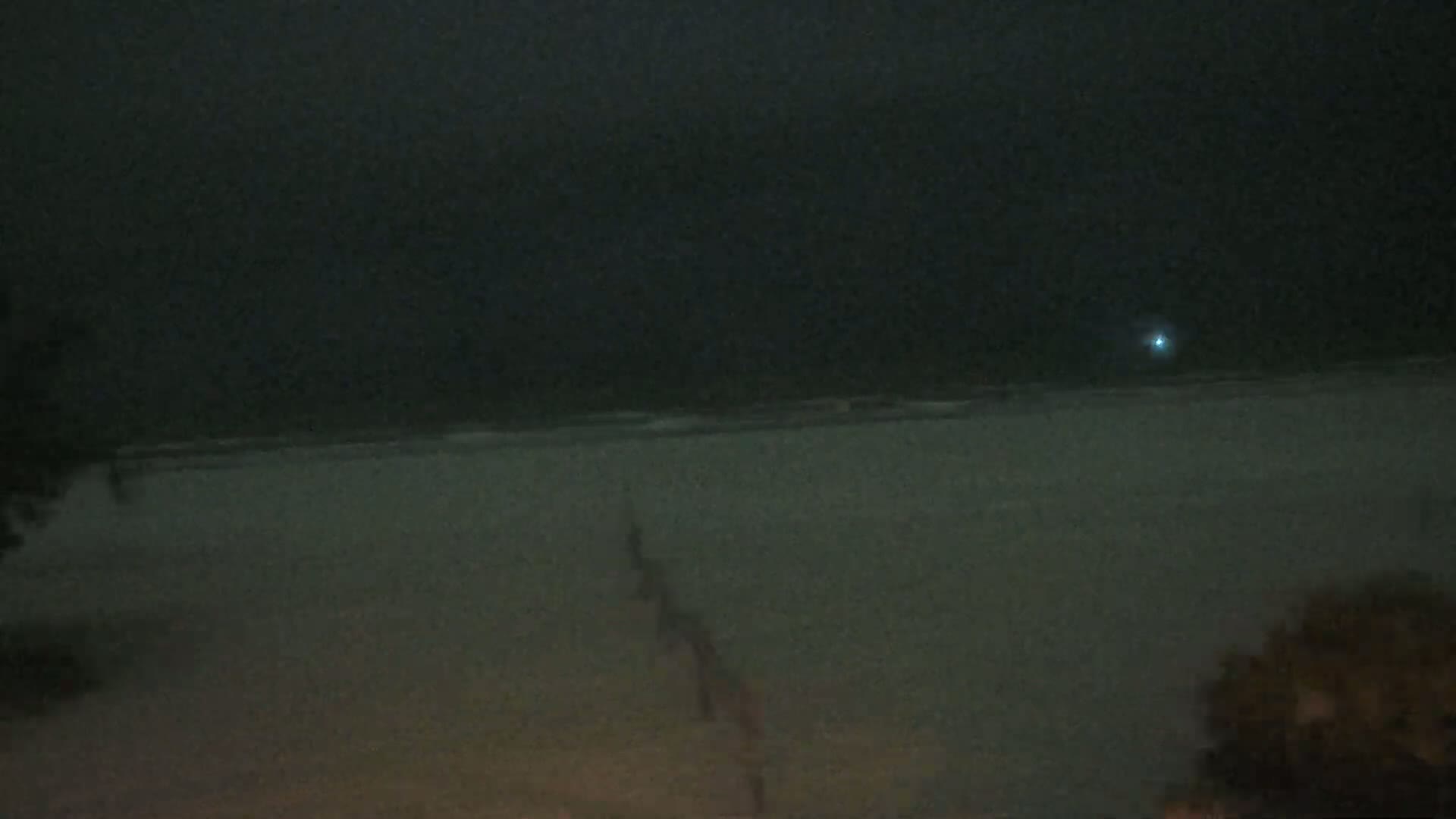 A dark, nighttime beach scene shows a mostly calm ocean under a cloudy sky, with vegetation visible in the bottom-right and bottom-left corners.