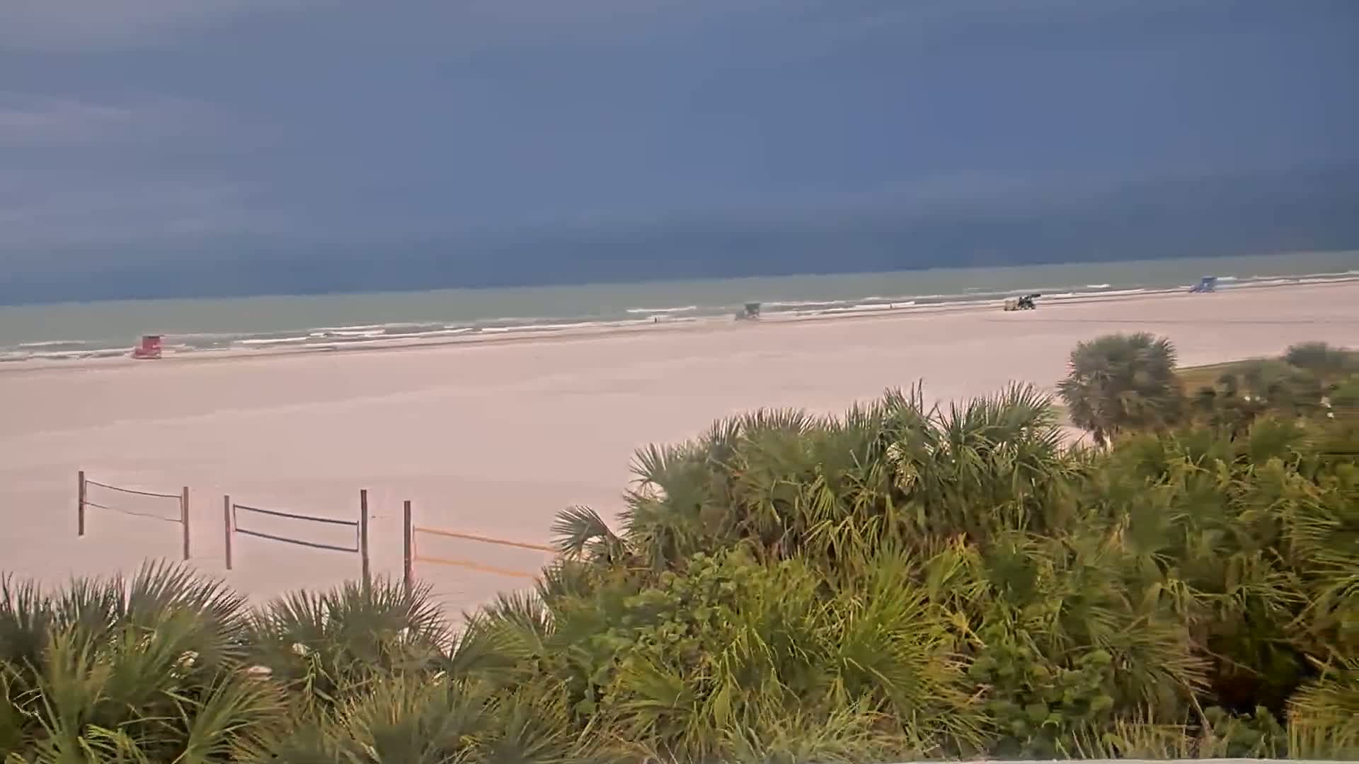 A wide sandy beach with calm waves under an overcast sky is partially obscured by tropical vegetation in the foreground.
