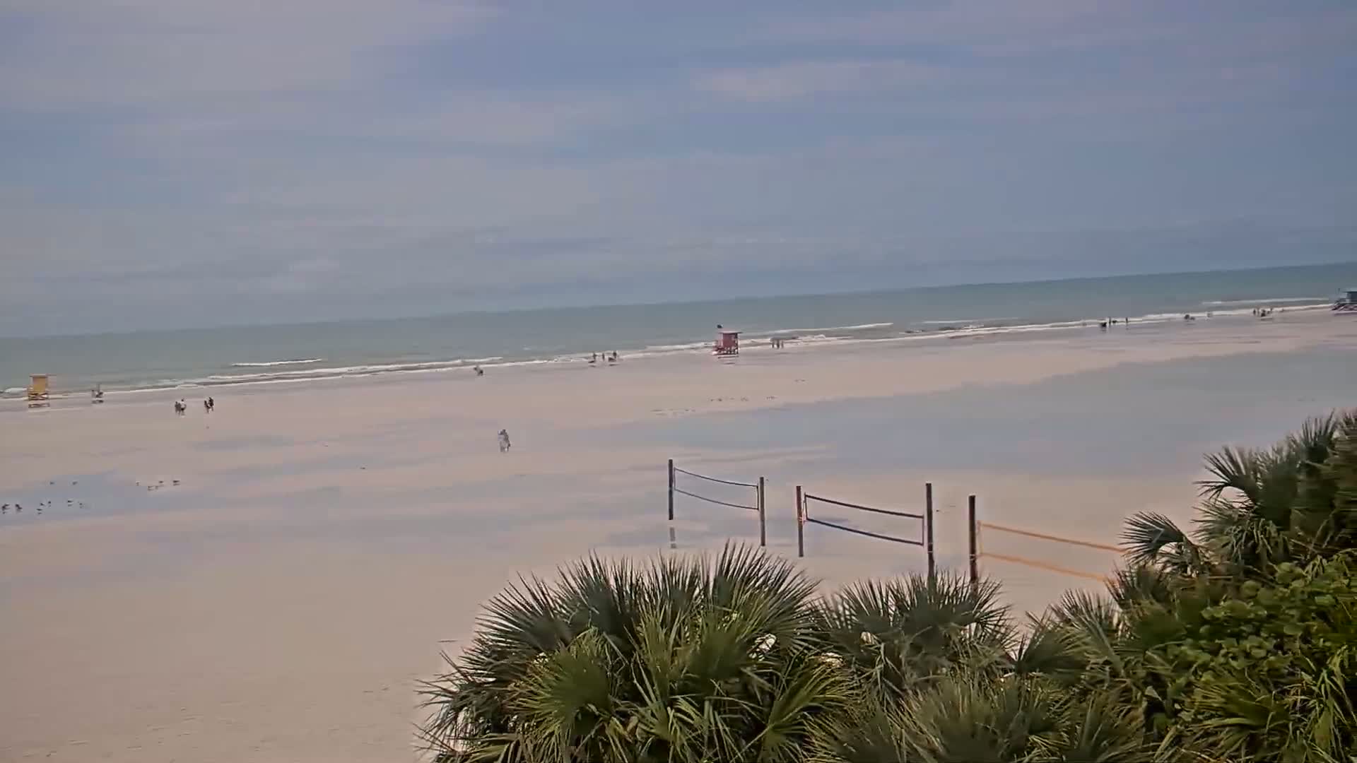 A mostly empty beach with a lifeguard stand, volleyball nets, and a few people visible under an overcast sky.