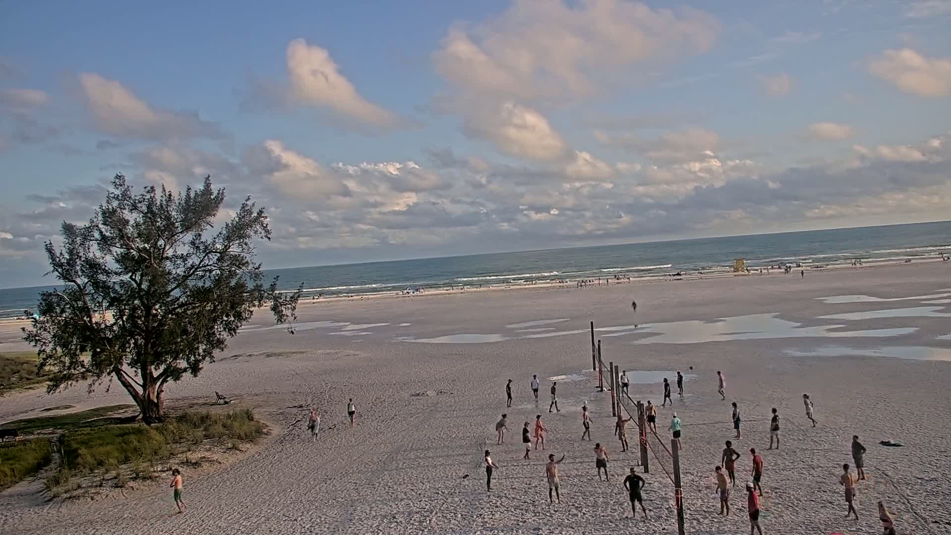 A wide shot of a beach with a volleyball game in progress under a partly cloudy sky, with several small puddles on the sand near the water's edge.