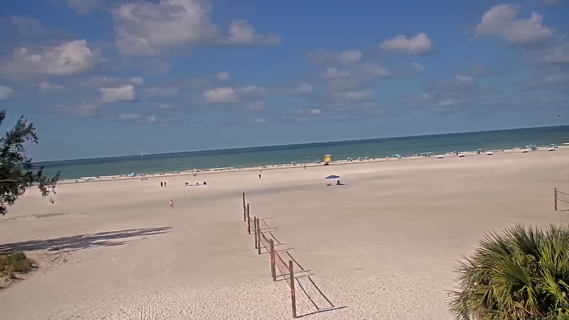 A mostly sunny day at a beach shows a wide expanse of light-tan sand with scattered people, a volleyball net, and a calm ocean in the background.