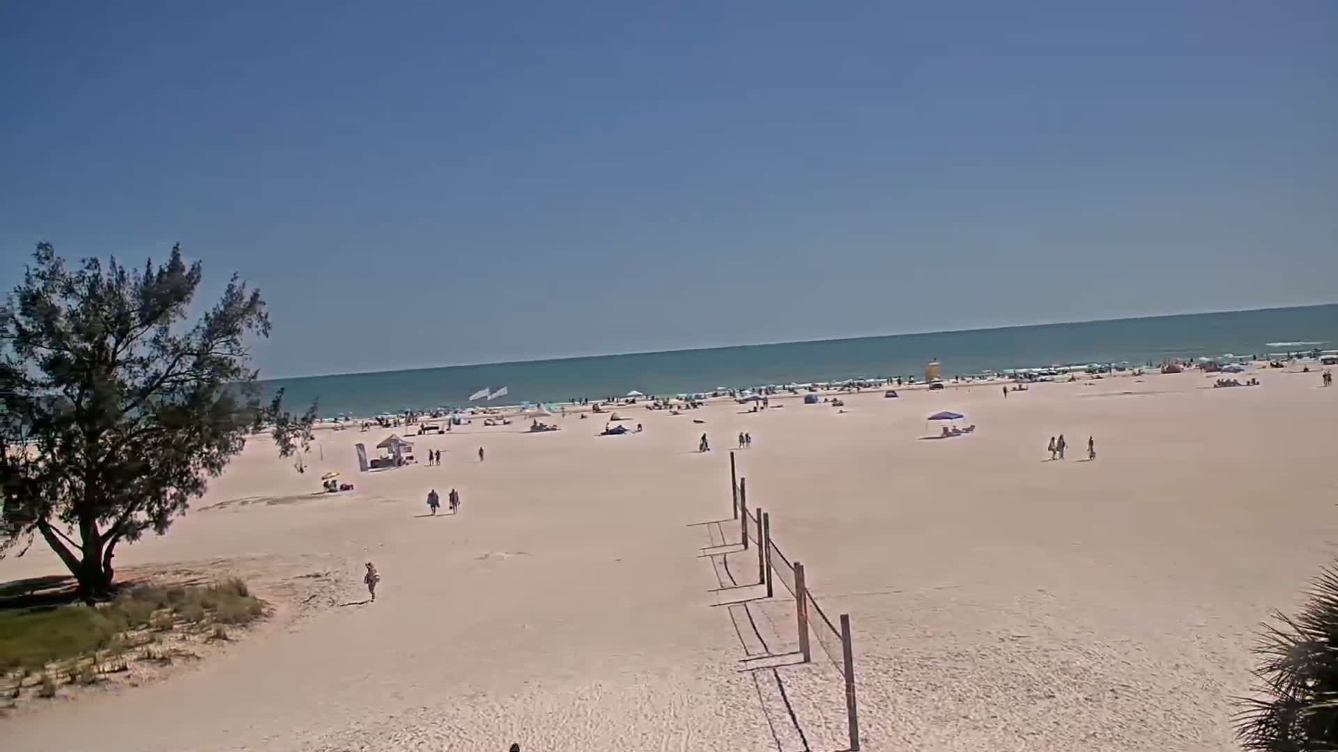 A mostly sunny day at a beach shows a wide expanse of light-colored sand with scattered people, umbrellas, and a volleyball net, all extending to a calm ocean under a clear blue sky.