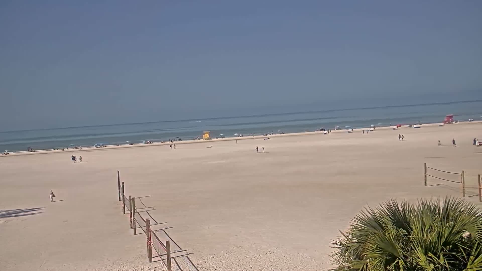 A mostly empty, wide sandy beach stretches to a calm ocean under a clear blue sky.